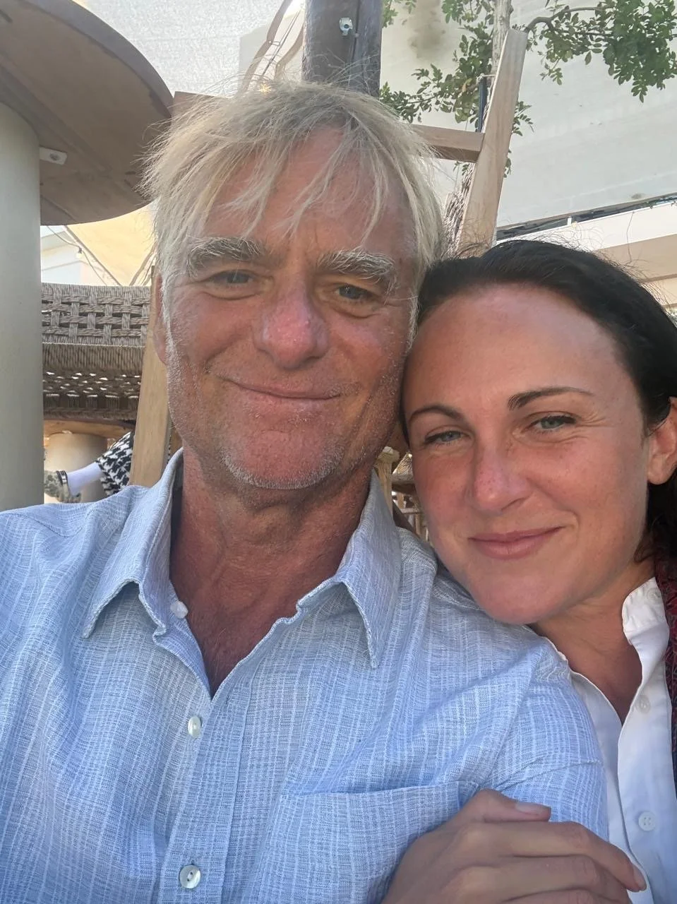 A man and a woman smiling closely for a selfie outdoors. The man has white hair, facial hair, and is wearing a light blue shirt. The woman has dark hair, is wearing a white blouse, and is resting her head on the man's shoulder.
