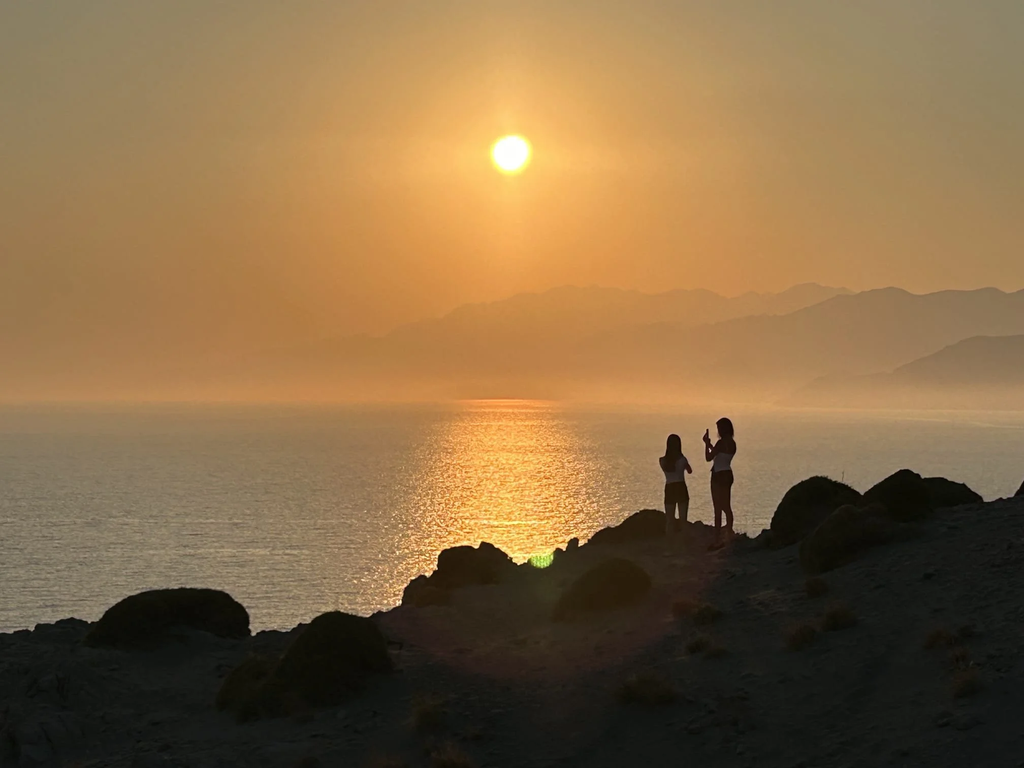 Two people standing on rocky coastline at sunset, with the sun reflecting on the ocean and mountains in the background.