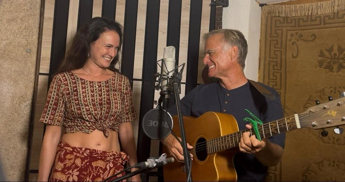A woman and a man smiling at each other. The man is playing an acoustic guitar and they are in a music studio setting.