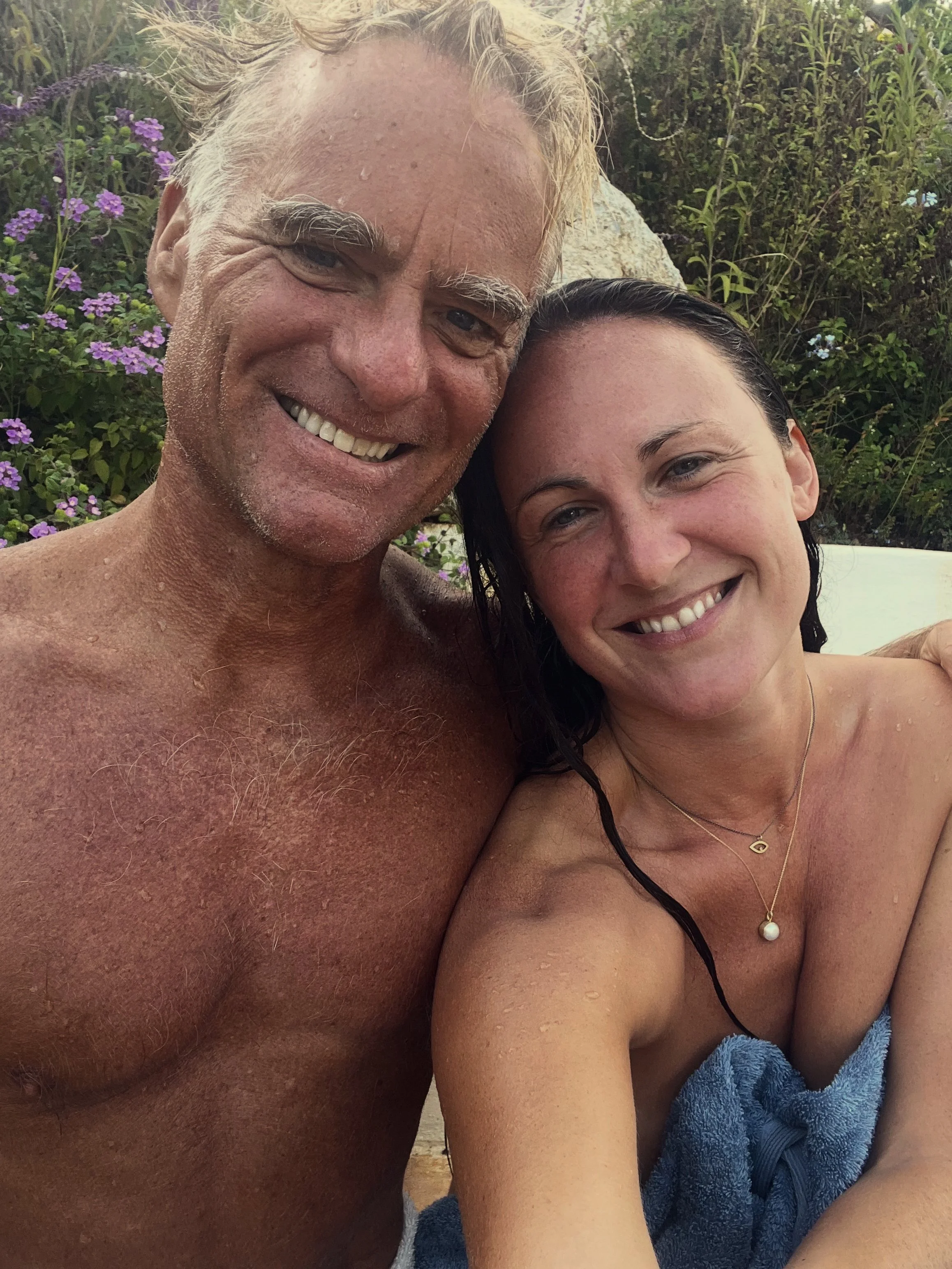A man and woman smiling close together outdoors with purple flowers and greenery in the background.