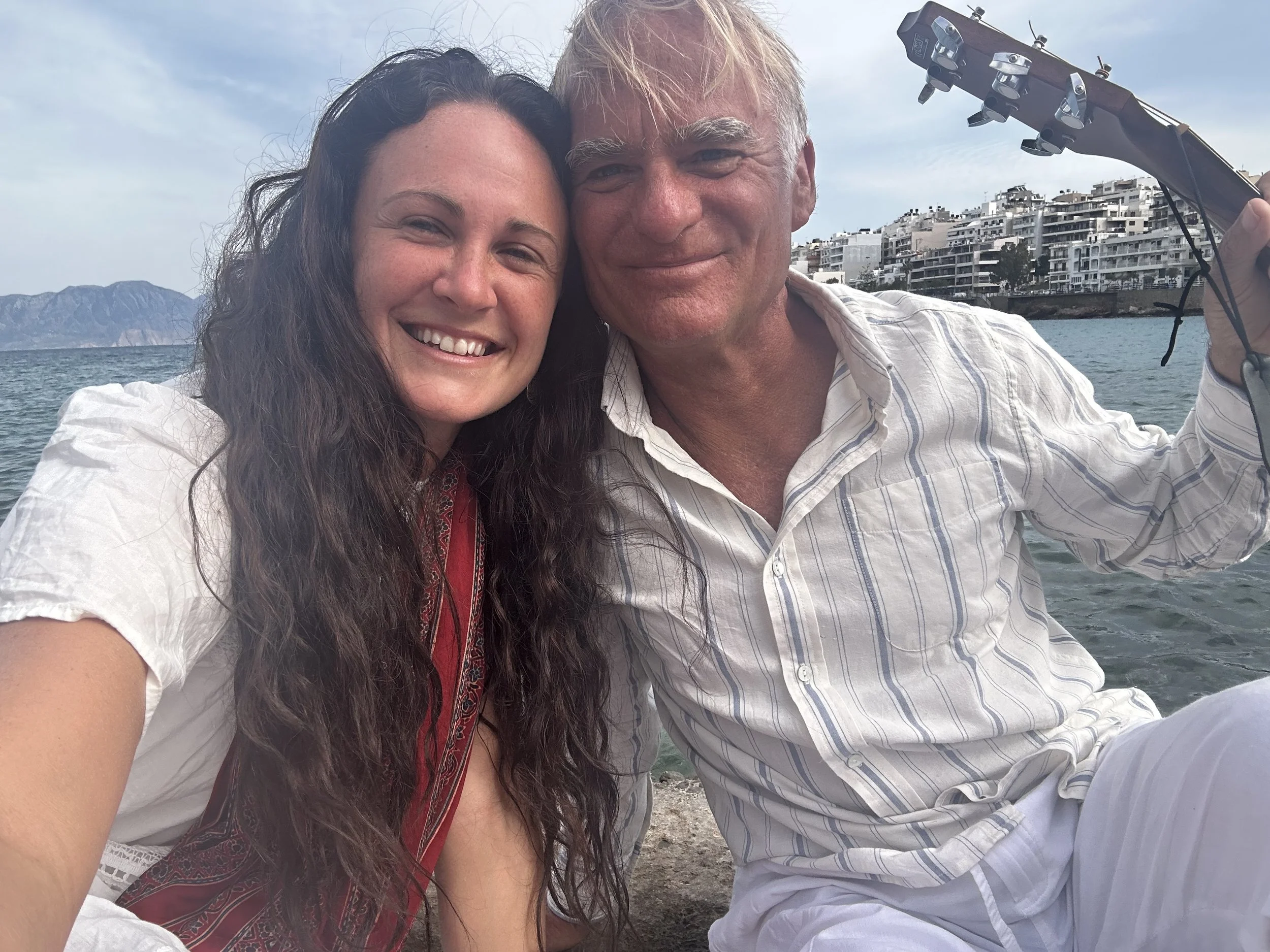 Close-up of a smiling woman with long, dark, curly hair and a smiling man with short, blonde hair taking a selfie together outdoors near water with a cityscape and mountains in the background.