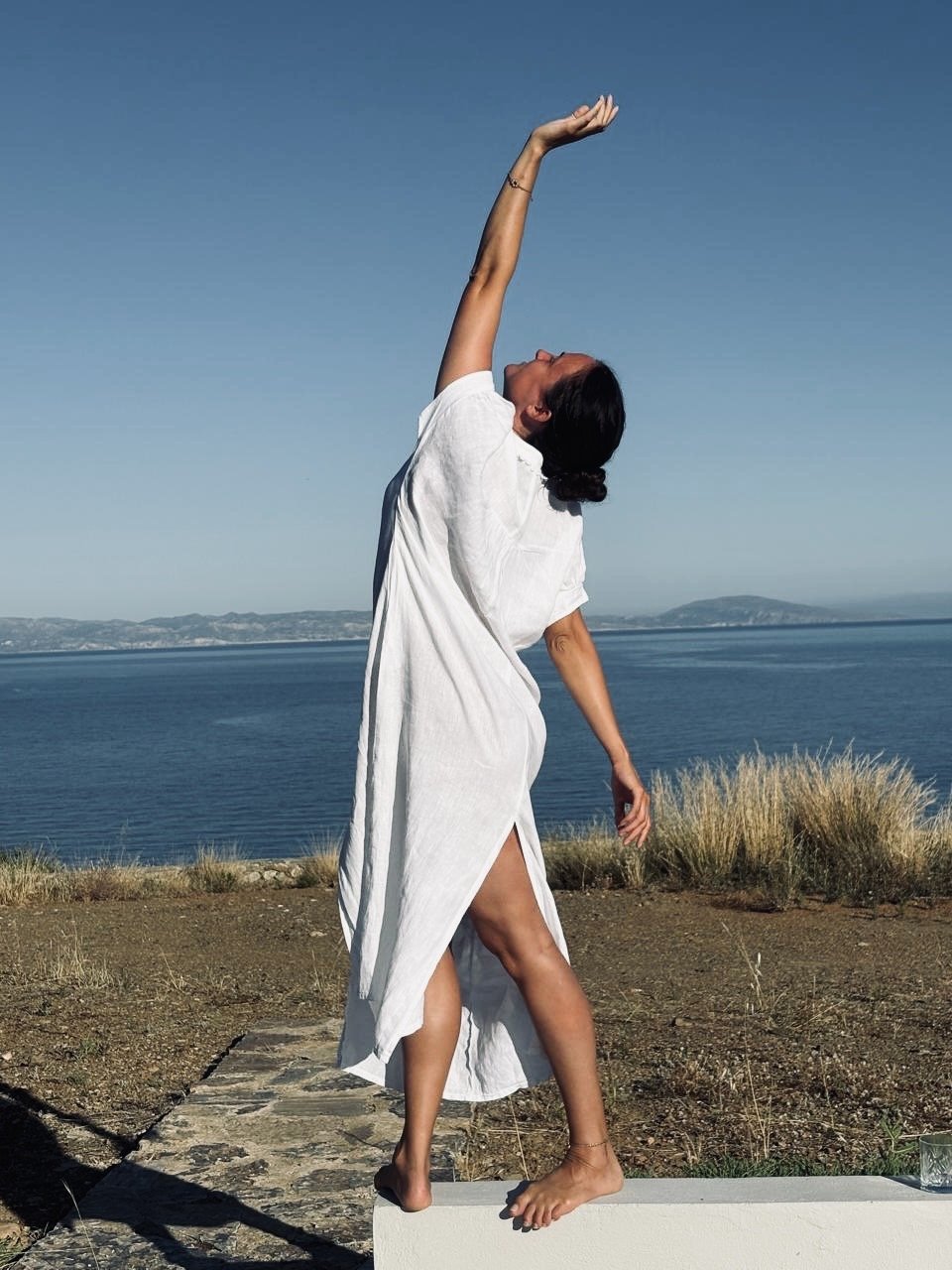 A woman in a white dress standing outdoors on a stone platform near a body of water, stretching her arm upward with a clear sky and distant land in the background.