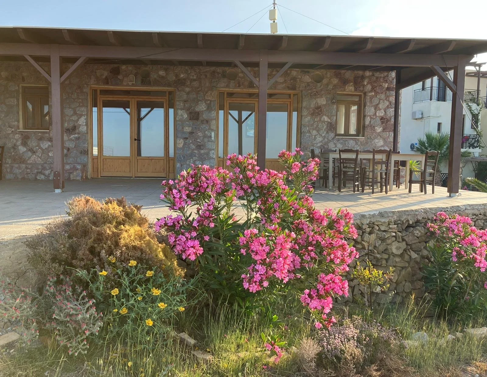 Stone house with patio, wooden door and windows, surrounded by pink and yellow flowers, with outdoor dining table and chairs.