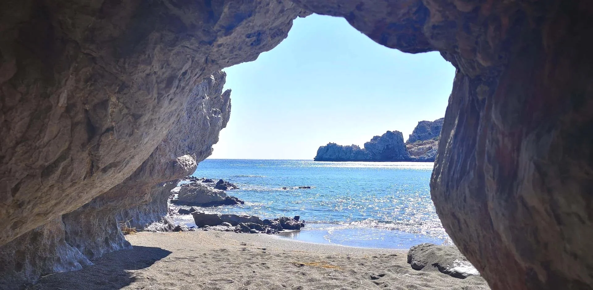 View of a beach seen through a large rock arch, with the ocean, rocks, and clear sky in the background.