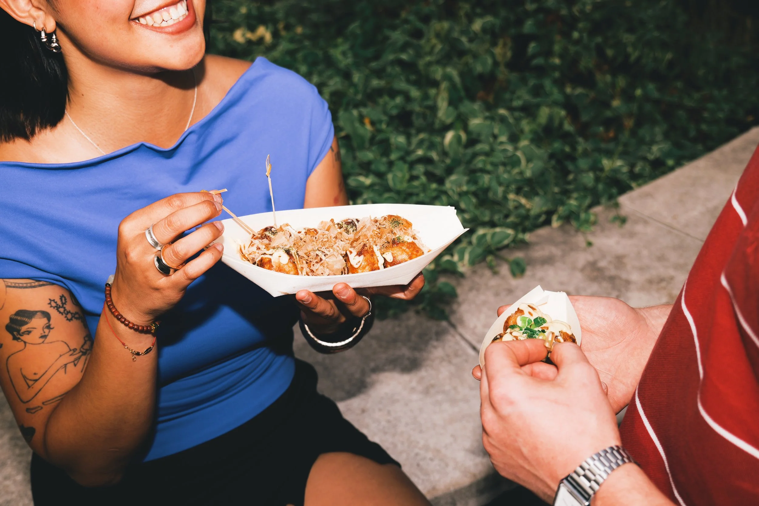 Close-up of two people exchanging plates of food outdoors. One person holds a white plate with Japanese takoyaki balls topped with sauce and bonito flakes, smiling, while the other holds a small plate with toppings in London.