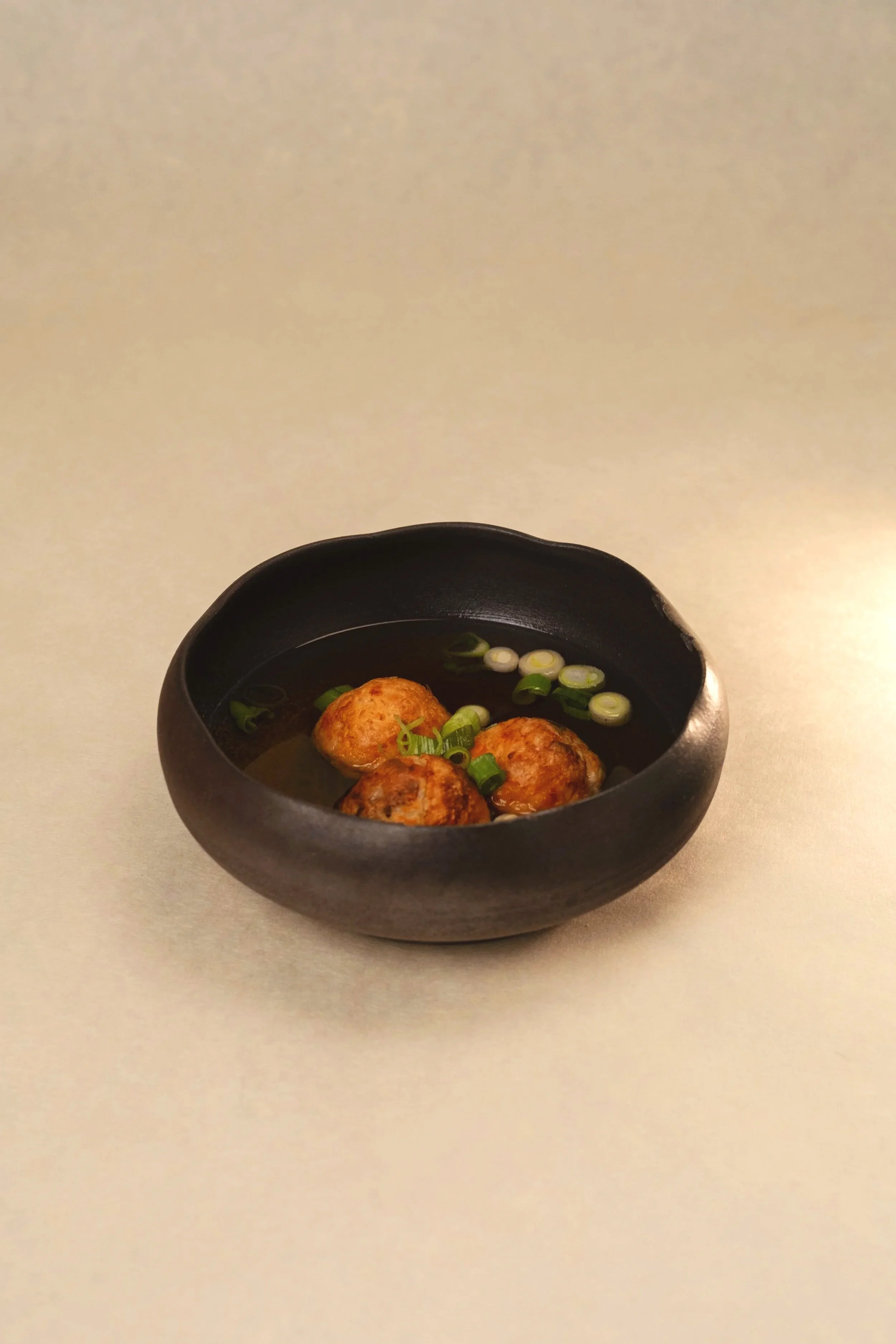 A black bowl with a few Japanese meatballs (tsukune) garnished with chopped green onions, served in a sauce. Takoyaki Soup broth.