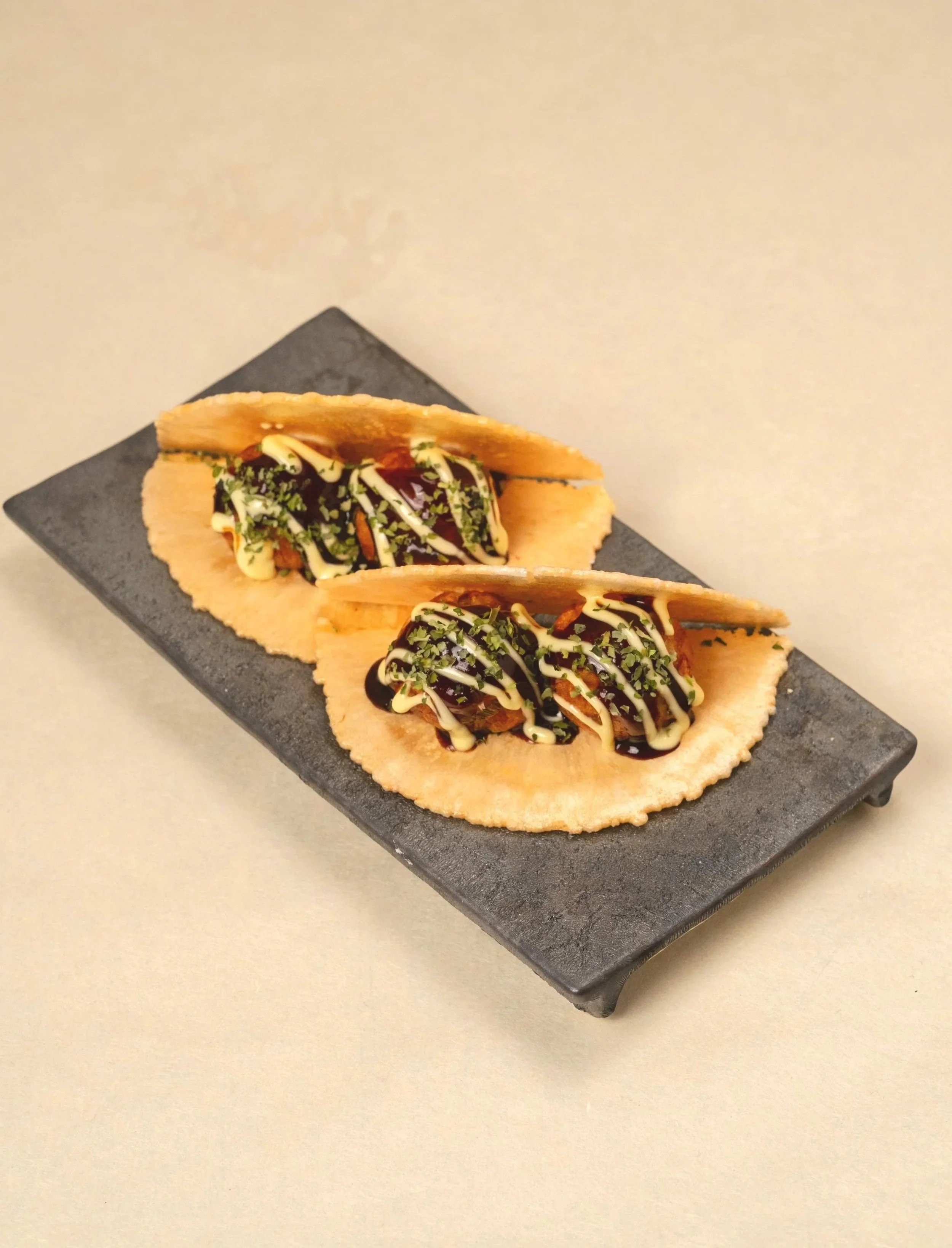 Three tacos filled with meat, topped with chopped herbs, shredded cheese, and drizzled with dark sauce and light sauce, served on a black rectangular plate. Takosen. Taco-sen Sando.