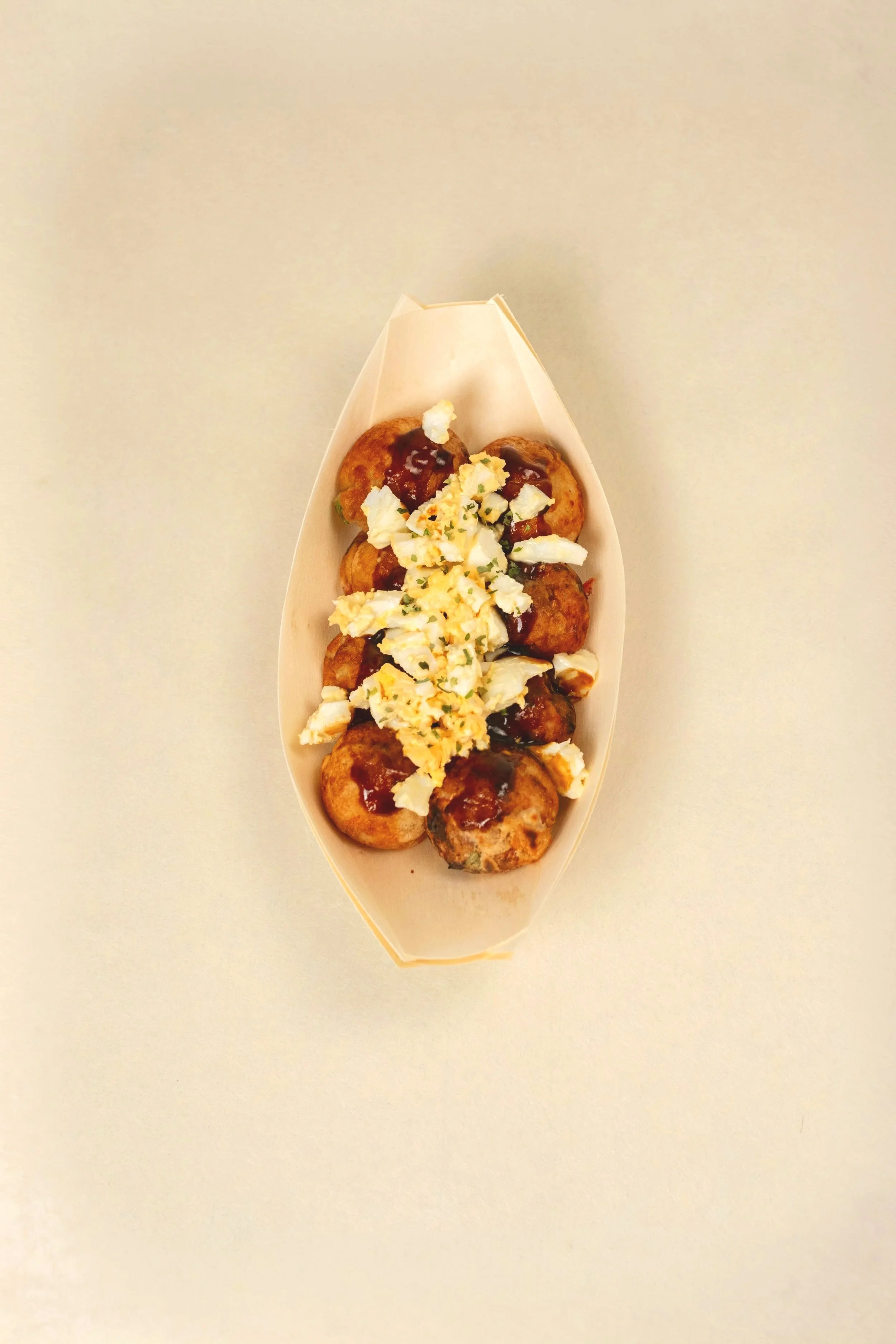 A boat-shaped tray with takoyaki topped with sauces, mayonnaise, and chopped green onions on a plain light-colored background. Egg salad.