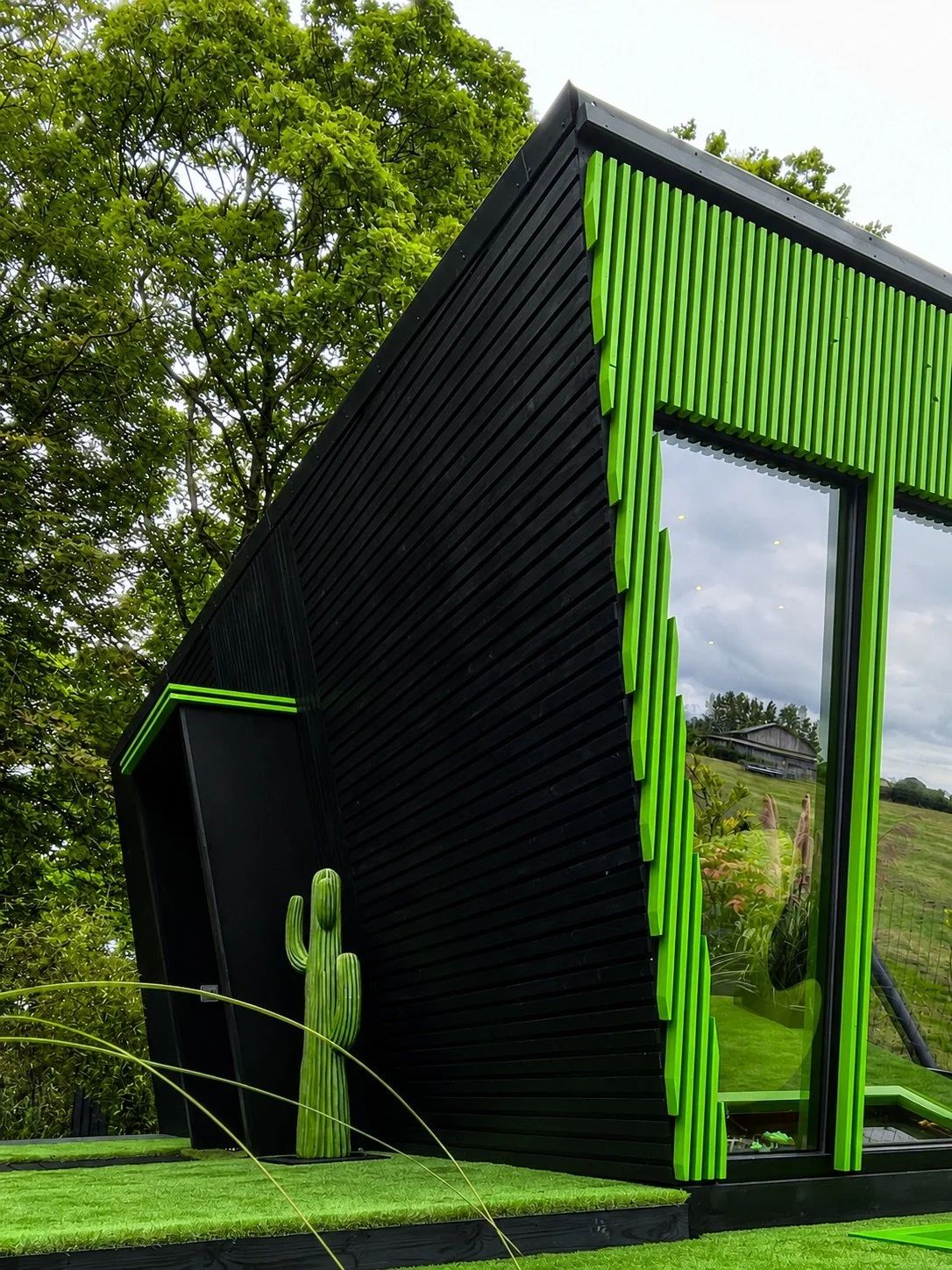 Low-angle view of NOCTURNE showing its black angular exterior, vivid green trim and glazed side beneath mature trees.