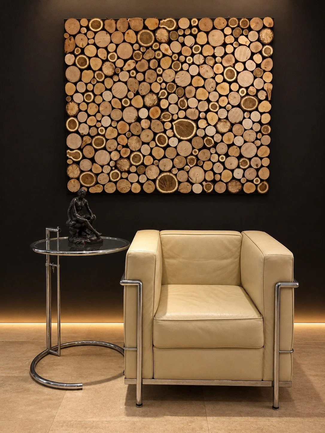 Square wall artwork made from natural timber rounds above a cream leather and chrome armchair against a dark wall.