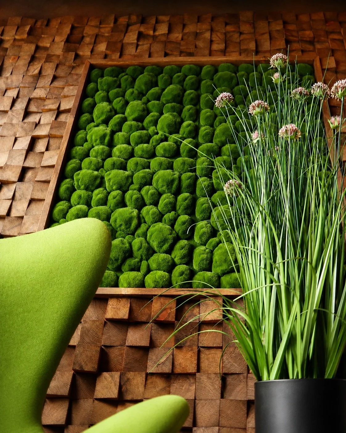 Close-up of a modern interior wall decorated with inset green moss balls, surrounded by wooden panels and green houseplants, with a lime green chair partially visible in the foreground.