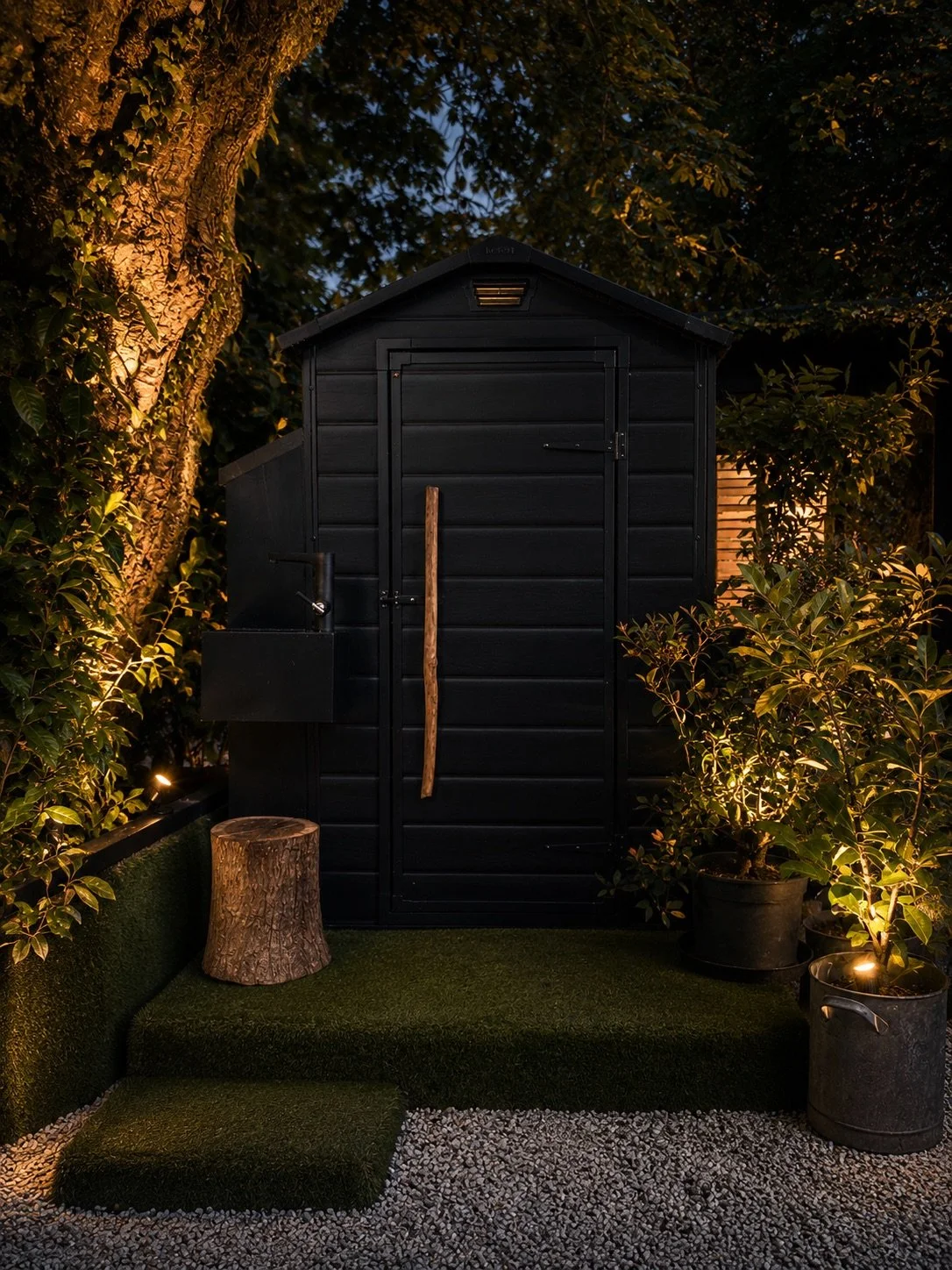 Compact black timber outhouse with an oversized natural-wood door handle, surrounded by softly illuminated planting.