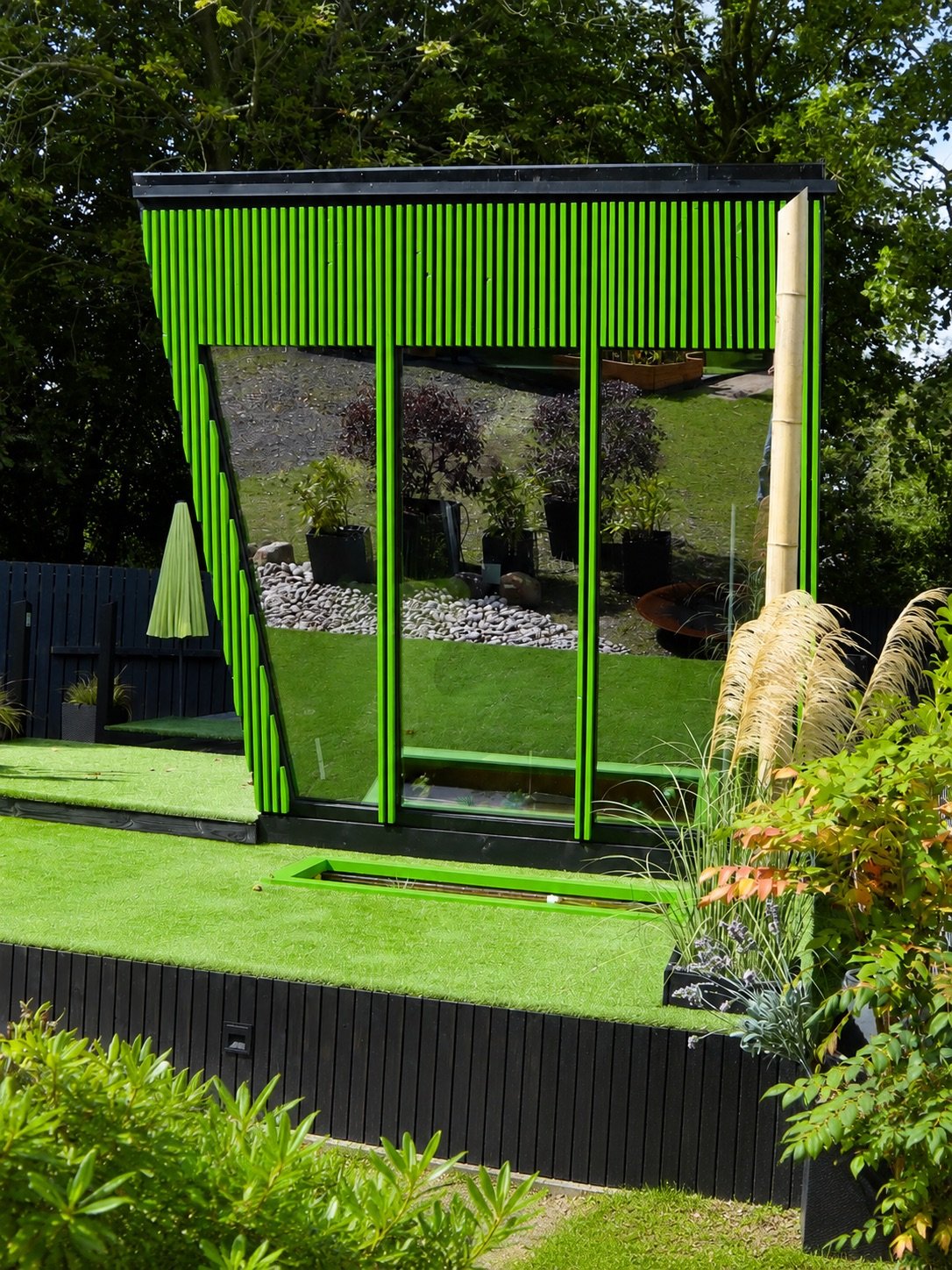 Front view of NOCTURNE showing its glazed end wall, vivid green framing, black cladding and raised timber deck.