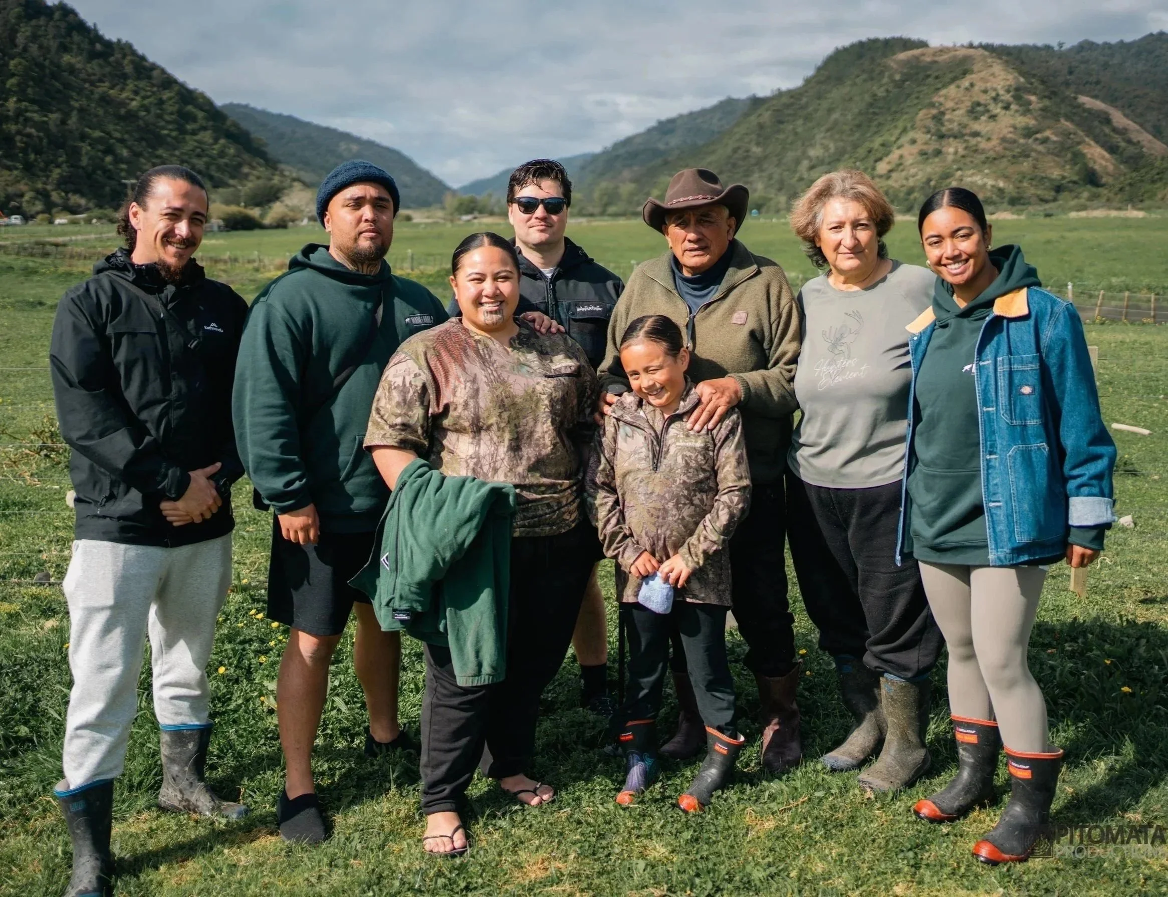 Group of eight people outdoors in a grassy field with mountains in the background, some wearing camouflage or outdoor clothing, standing together for a photo.