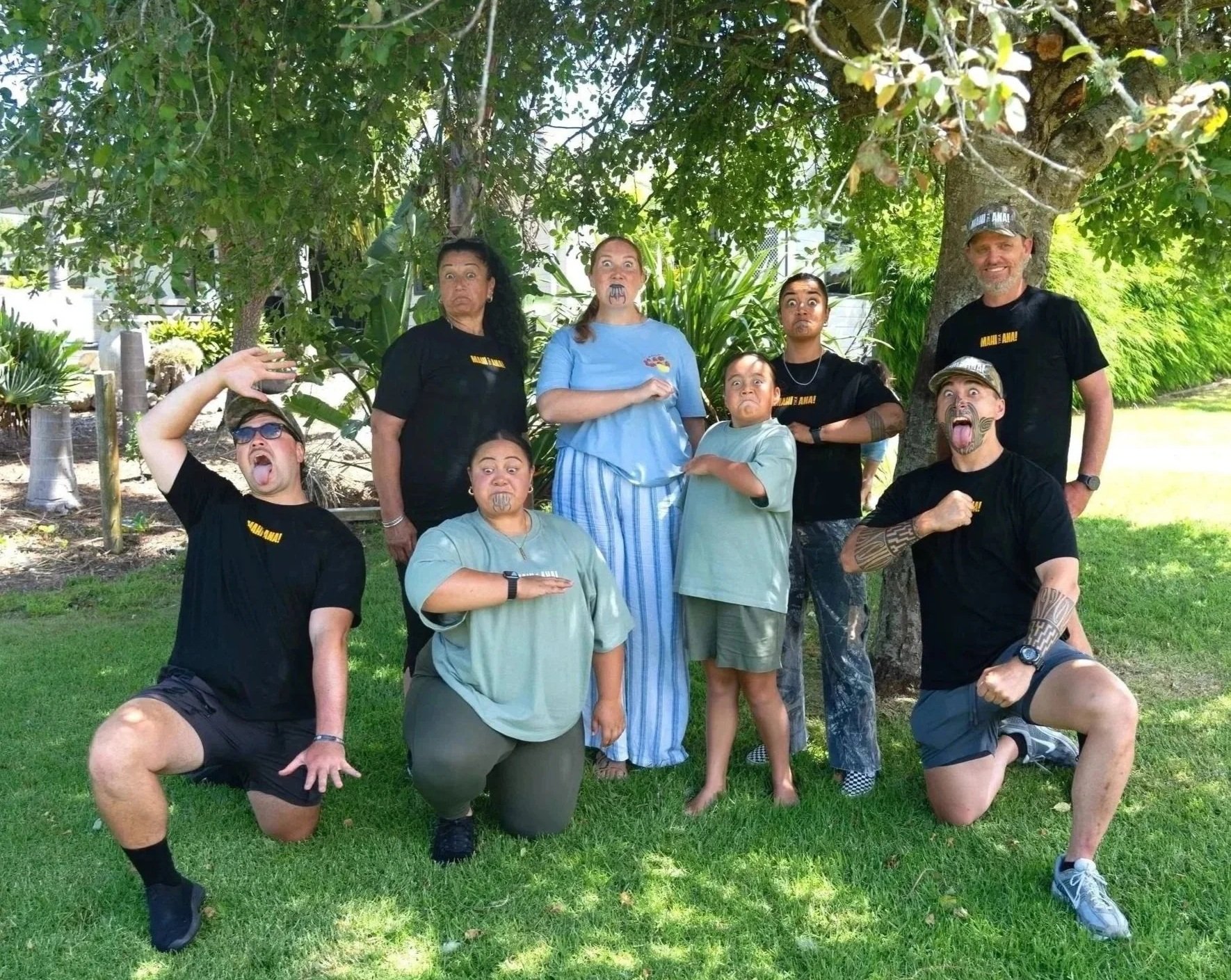 Group of eight people outdoors posing playfully with facial expressions and gestures in front of a tree and greenery.