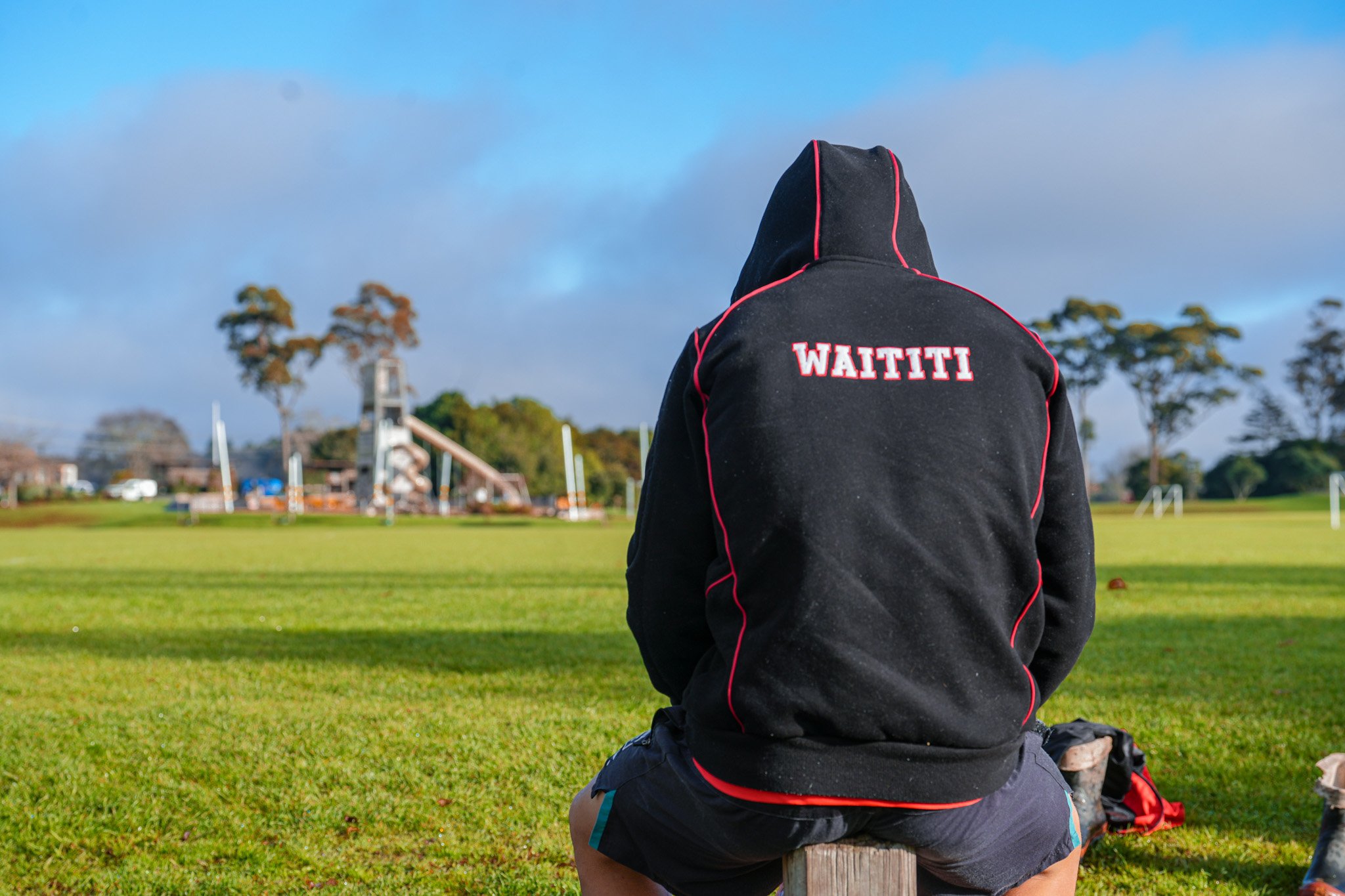 A person wearing a black hoodie with red trim and the word "WAITITI" on the back, kneeling on a wooden post in a grassy field, with trees and a playground in the background under a blue sky with some clouds.