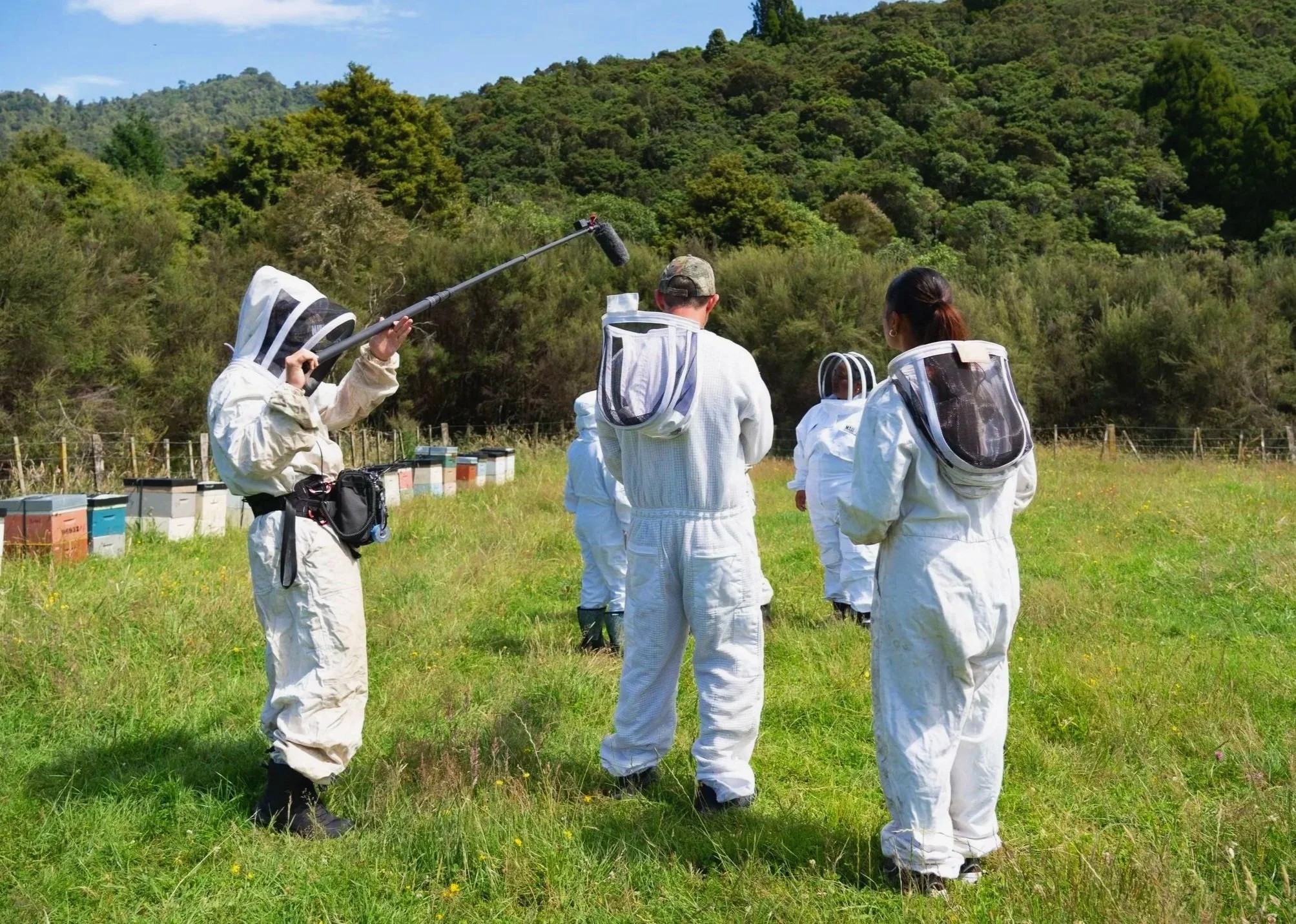 Group of people in protective suits standing in a grassy field, surrounded by trees and forest, with blue sky overhead.