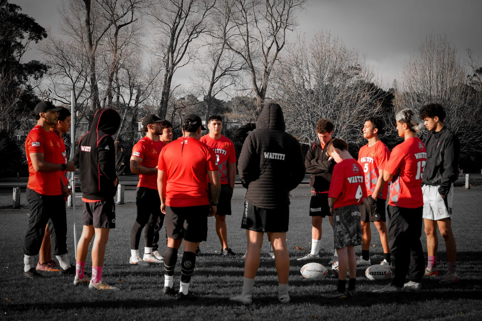 A group of young rugby players in red sports uniforms gathered around their coach on a field with leafless trees and cloudy sky in the background.