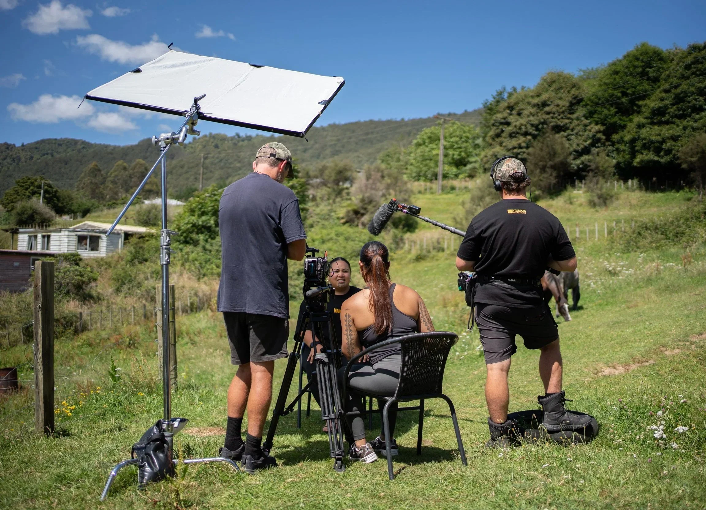 Filmmakers on a grassy hill filming a woman sitting on a chair, with a large reflector and camera equipment under a clear blue sky with scattered clouds and green hills in the background.