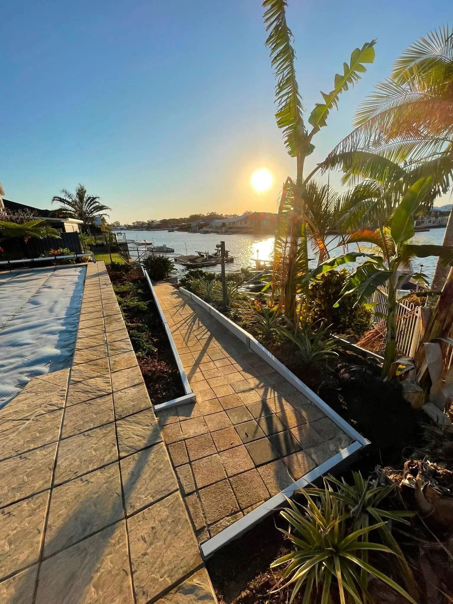 Sunset over a canal with boats, tropical plants, and a tiled patio in the foreground.