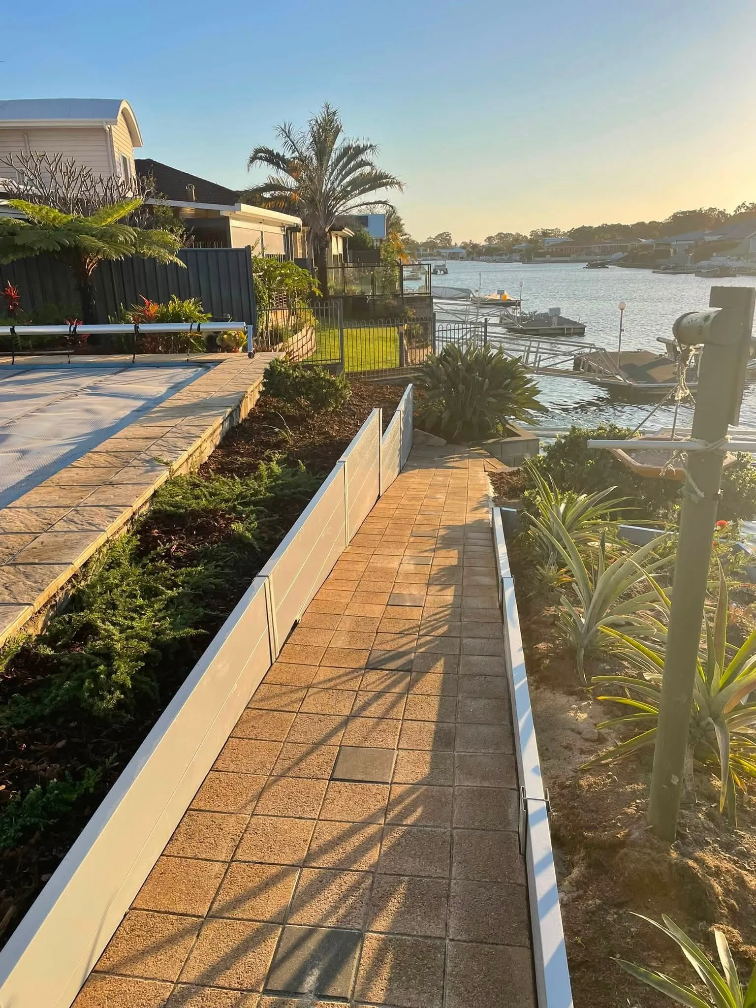 A backyard scene by a waterway during sunset, featuring a paved patio, lush plants, a black fence, a palm tree, and boats docked along the water.