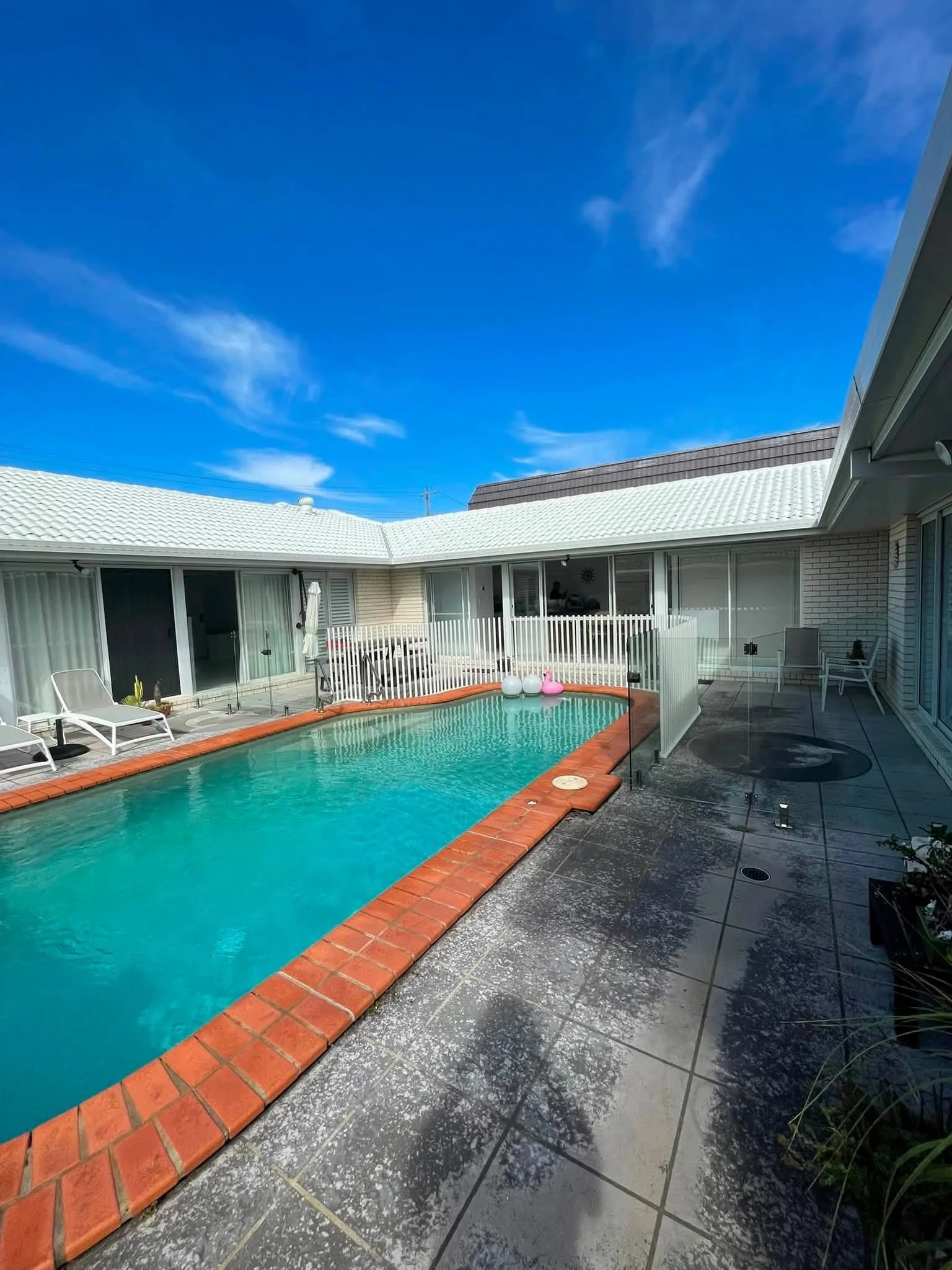 Backyard with a swimming pool, white patio chairs, umbrellas, and a white fence under a blue sky with some clouds.