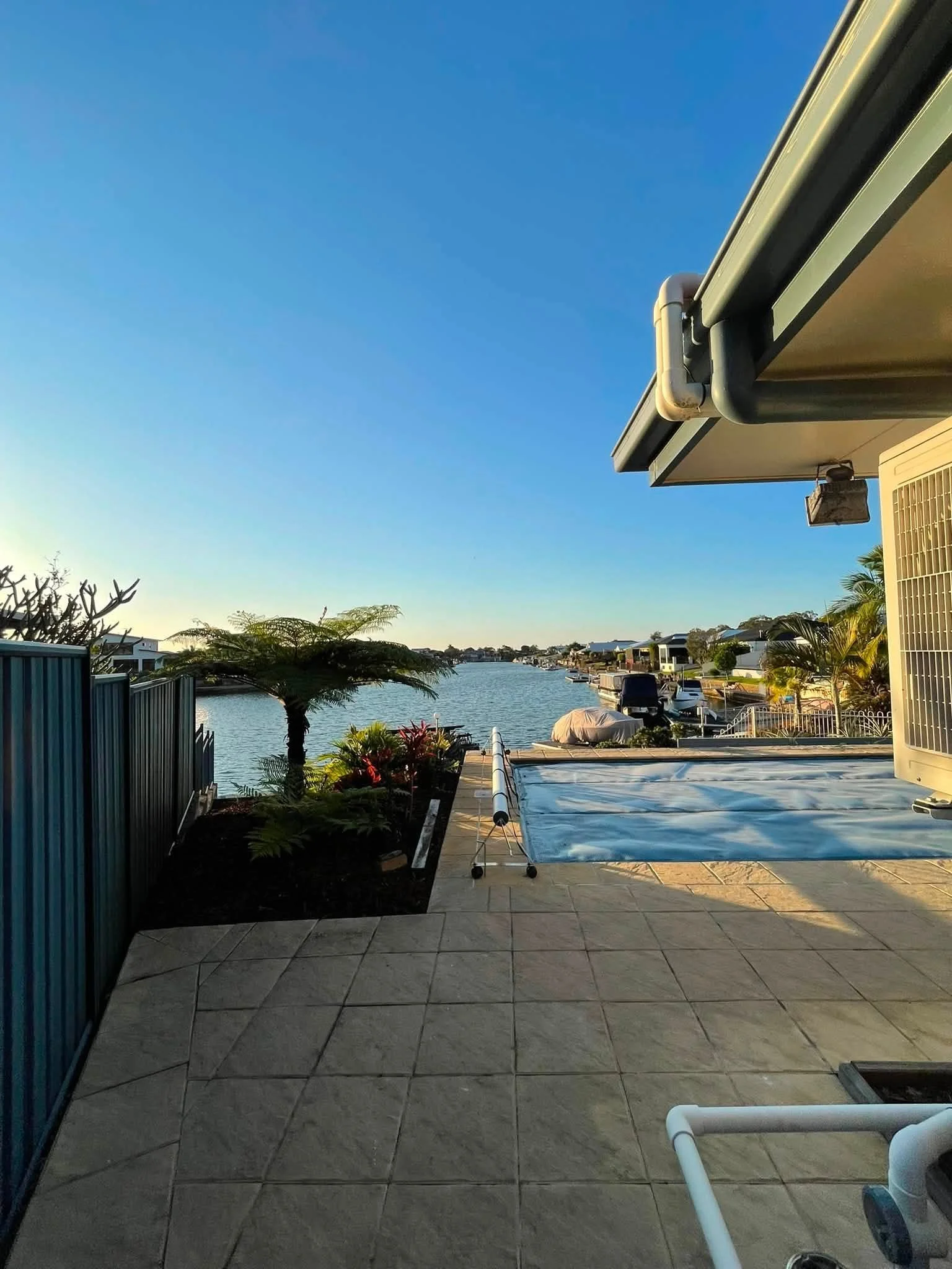 View of a waterfront backyard patio with potted plants, a small tree, outdoor furniture covered with a tarp, and a river with boats in the background under a clear blue sky.
