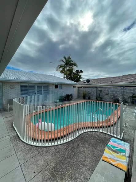 Residential backyard with an in-ground swimming pool, surrounded by a white safety fence, and a small colorful towel on a chair. The yard is enclosed by a brick wall, with a house and palm trees in the background under a cloudy sky.