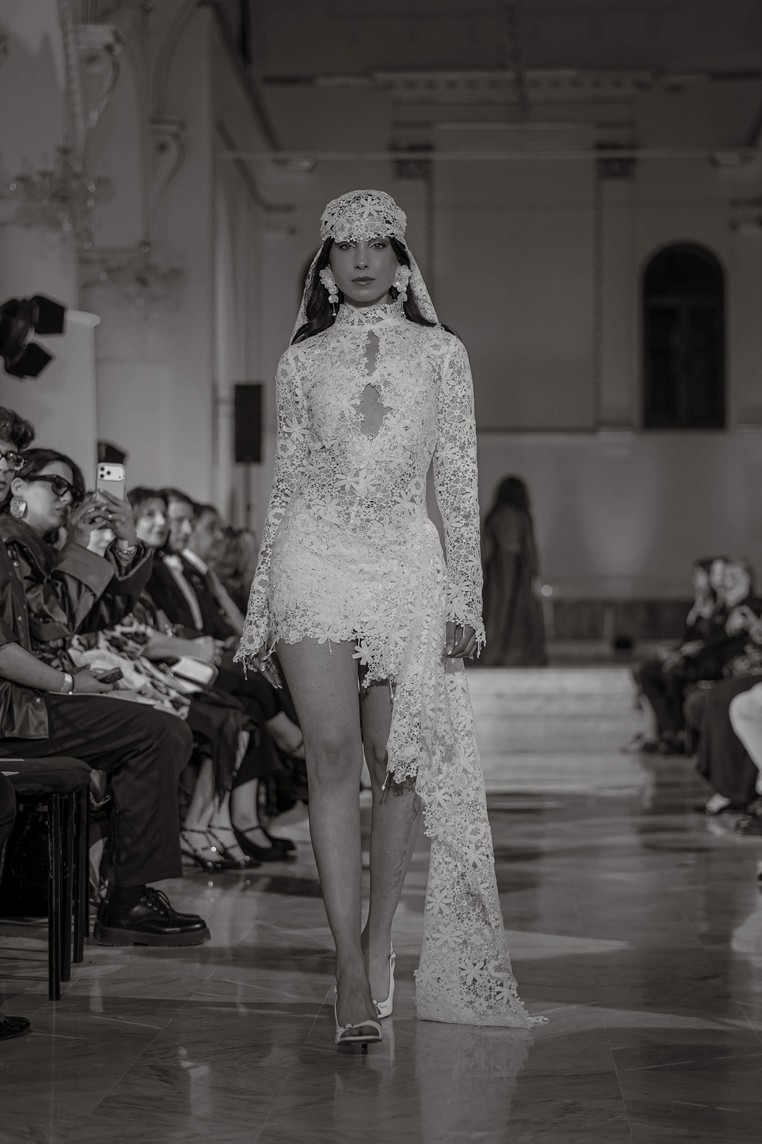 A female model walking down a runway wearing a lace wedding dress with a high slit, lace head covering, and statement earrings, while audience members are seated on either side, with some taking photos. Zsolea Bridal Gown