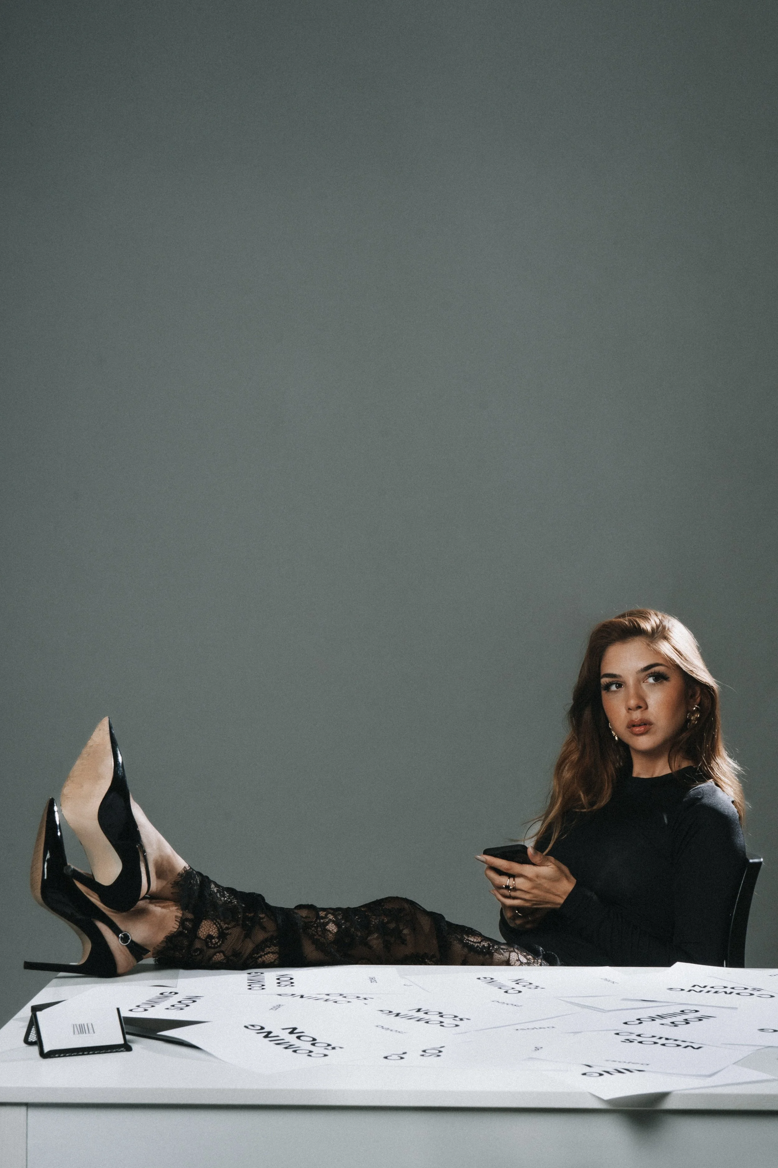 Woman with long, wavy, reddish-brown hair sitting on a table with her legs stretched out, wearing black lace high heels and a black dress, holding a phone, in a photo studio with gray background and a table covered with printed papers and a black notebook.