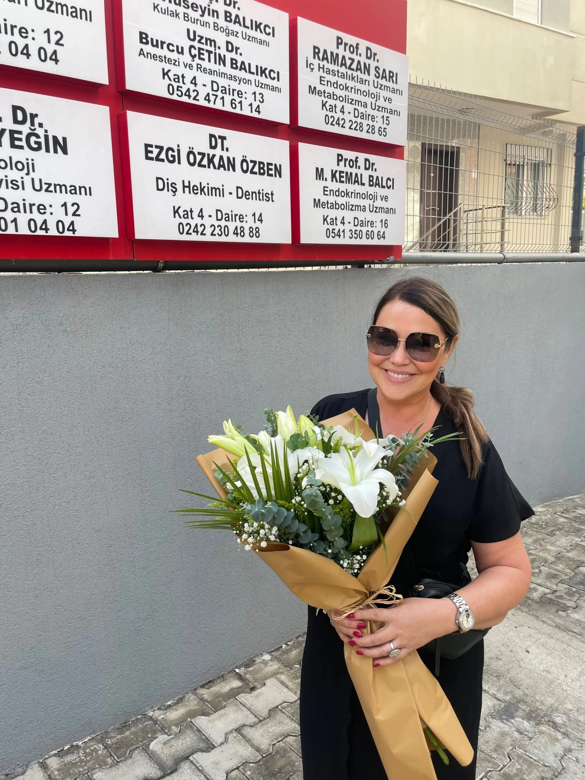 A woman wearing black clothing and sunglasses is smiling and holding a bouquet of white lilies and other white flowers wrapped in brown paper. She is standing outside in front of a gray wall and a sidewalk. In the background, there are several white and red signs with black text, possibly showing names and contact information of medical professionals.