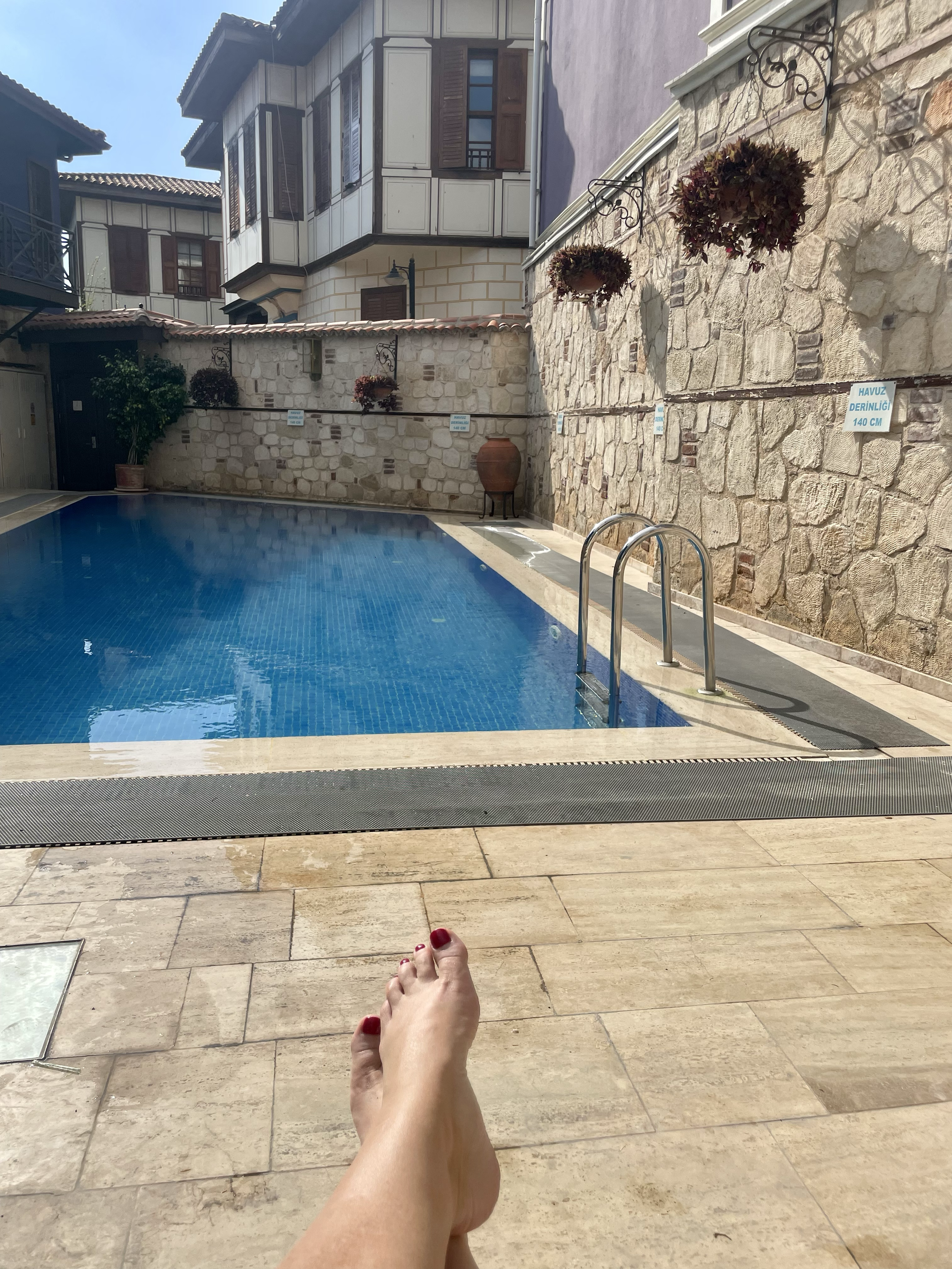 A relaxing outdoor swimming pool area with blue water, surrounded by stone tiles, with a person sunbathing and feet visible in the foreground. There are potted plants, stone walls, and a building with traditional Turkish architectural style in the ba