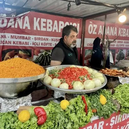 A man standing behind a food stall with a sign that reads 'DANA KEBAPÇISI' and other information in Turkish. On the table, there are large bowls of colorful ingredients including chopped tomatoes, onions, and shredded vegetables. In the foreground, t