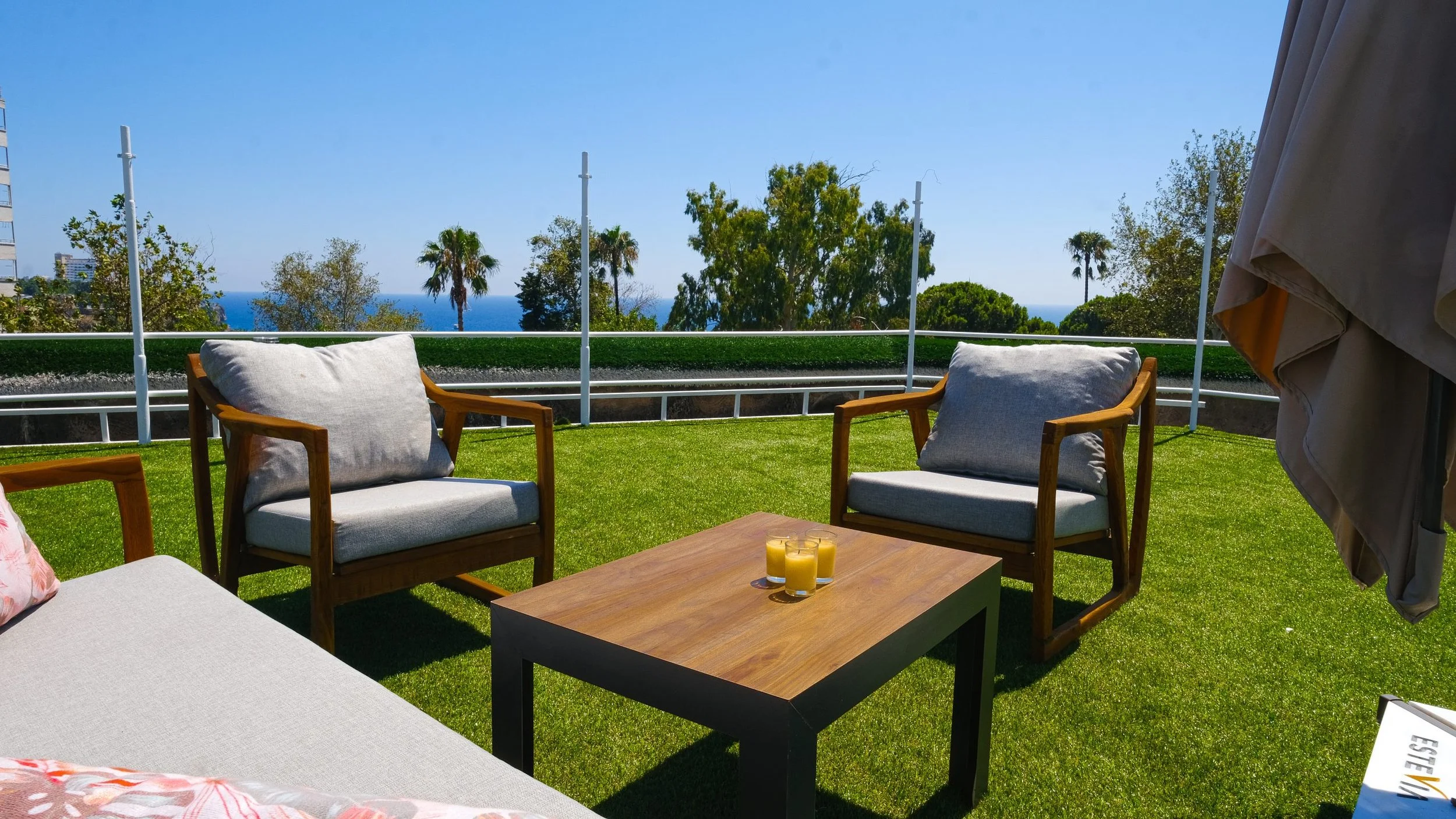 Balcony with outdoor furniture including chairs and a low table with candles, overlooking trees, palm trees, and the ocean under a clear blue sky.