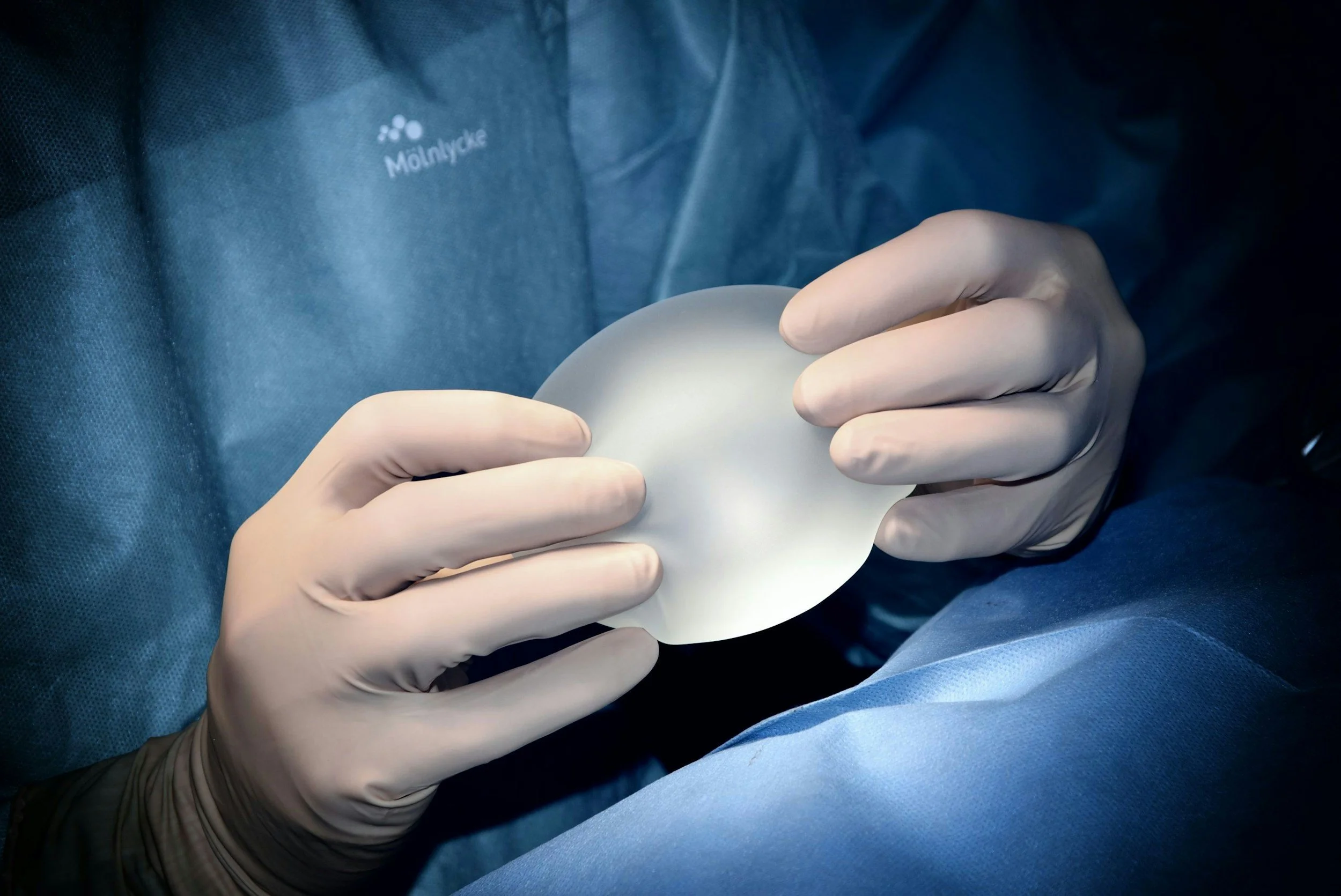 A healthcare professional wearing latex gloves holds a white implant or prosthetic device in a sterile environment.