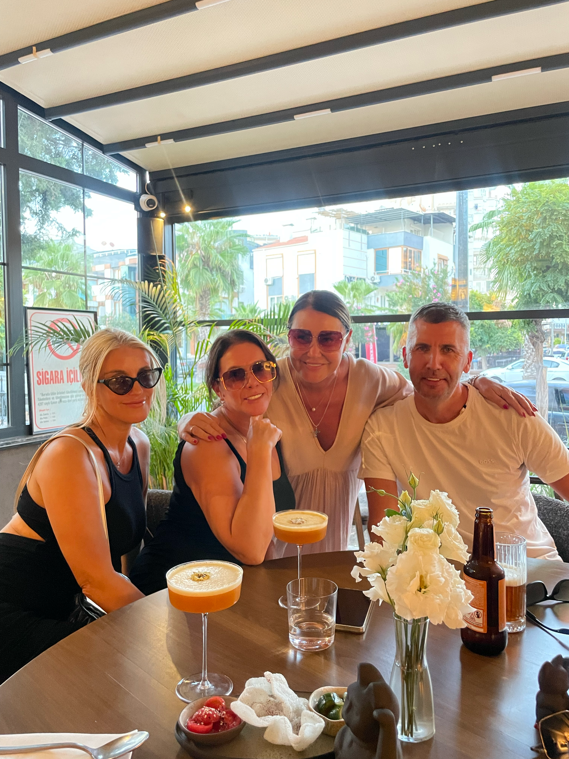 Four people sitting at a table in a restaurant, with drinks, flowers, and snacks, smiling and posing for the photo.