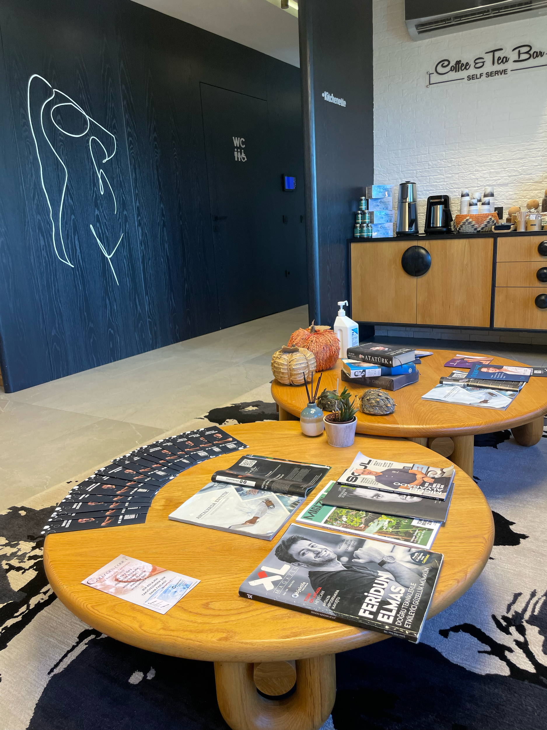 A round wooden table with magazines, pamphlets, and small decorative items such as a plant and pumpkins on top. Behind the table is a black wall with a sign that reads 'Coffee & Tea | Self-Serve' and a wooden cabinet with a coffee machine, thermoses,