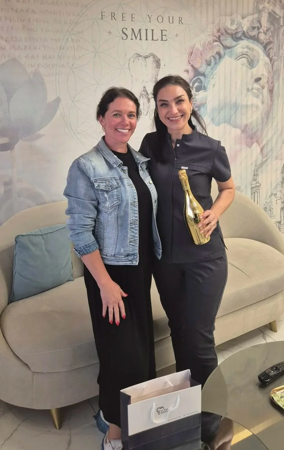 Two women standing close together, smiling, in a room with a mural that reads "Free Your Smile," with artistic faces and geometric patterns in the background. One woman is wearing a black outfit and holding a gold decorative bottle, while the other i