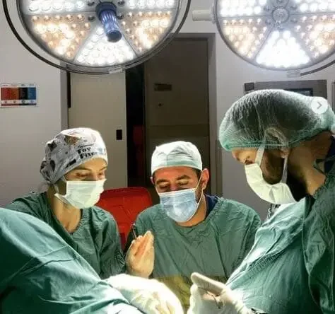 Four medical professionals in surgical scrubs, masks, and hair covers performing a surgery in an operating room under bright overhead lights.