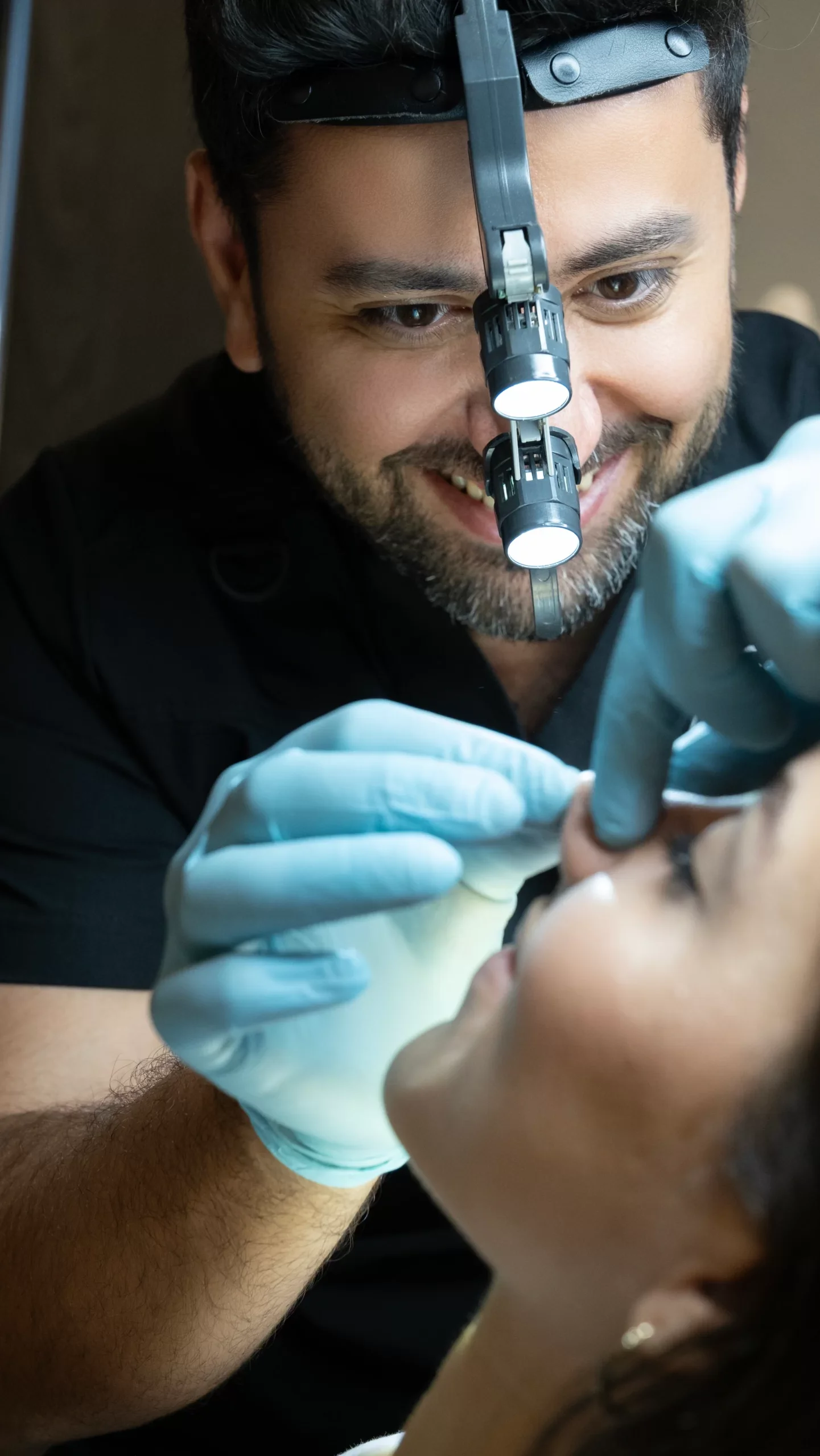 A male dentist or healthcare professional with a headlamp examining a patient's mouth. The patient is lying back with eyes closed, and the professional is wearing gloves, using dental tools for examination or procedure.