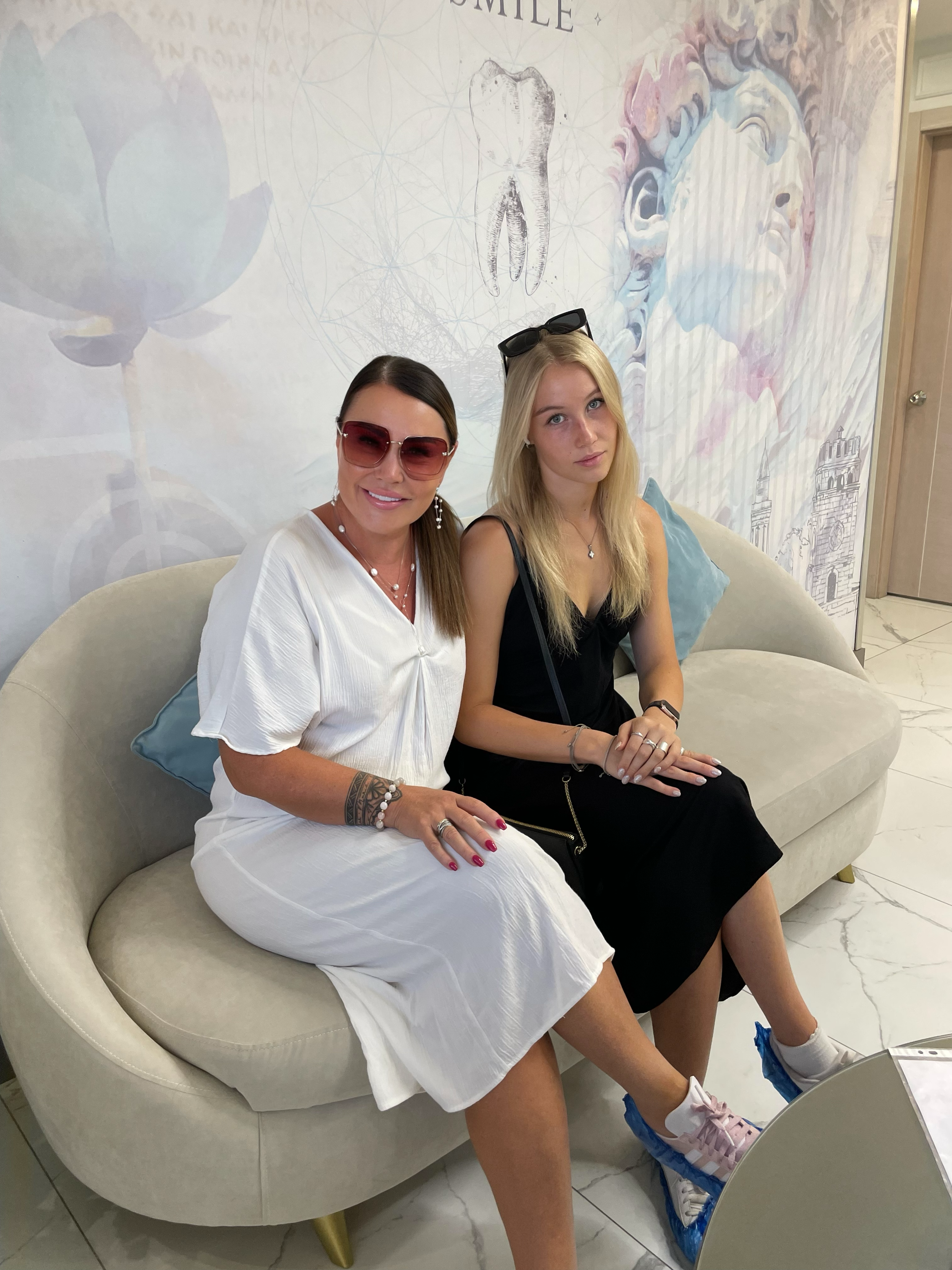 Two women sitting on a cream-colored sofa in a room with a light-colored marble floor. The woman on the left is wearing a white dress, sunglasses, and jewelry, and is smiling. The woman on the right has blonde hair, is wearing a black dress with a zi