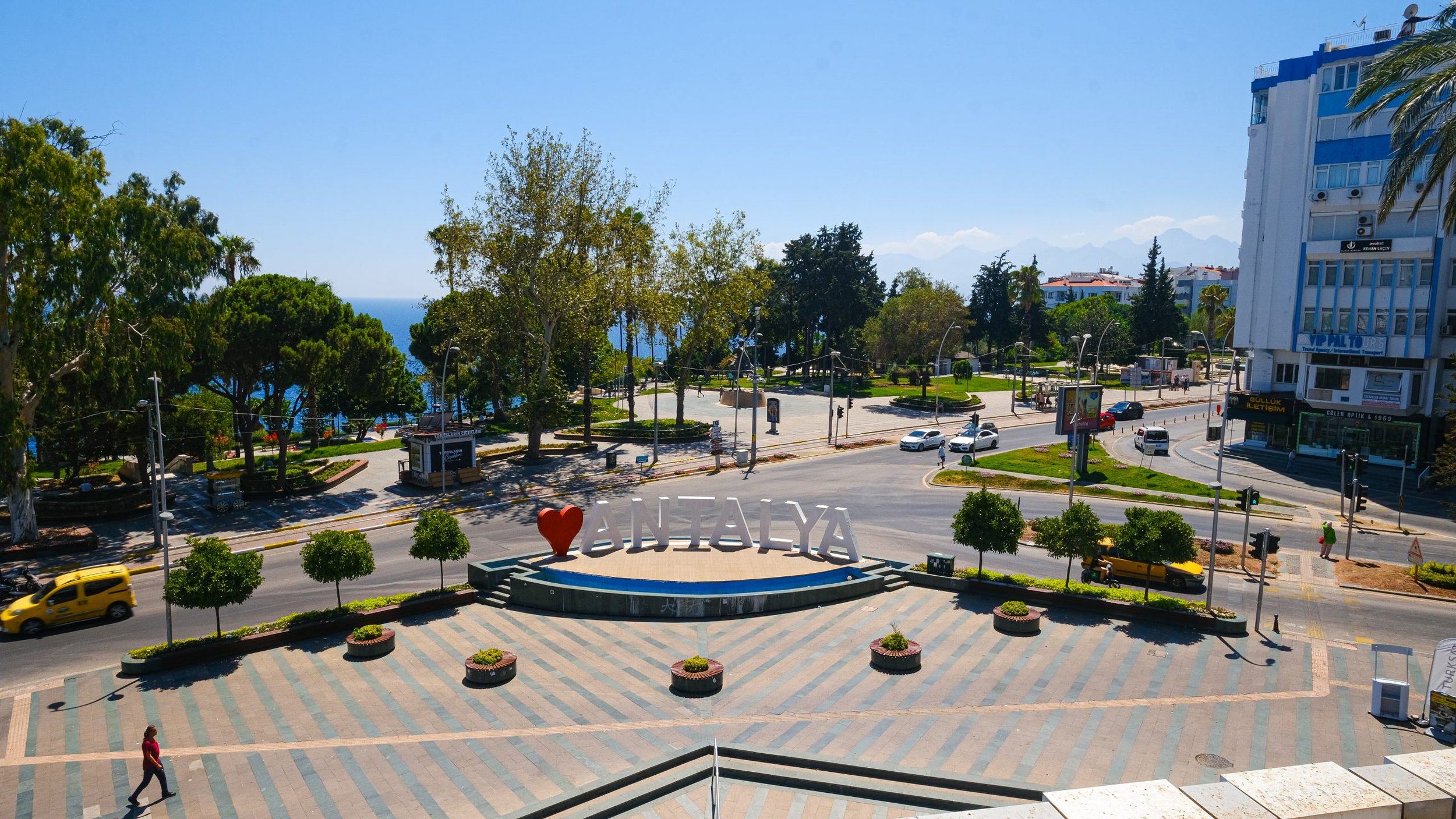 A city square with a prominent sign that reads "I love AntalyA" in front of a fountain, surrounded by trees, cars, and a few pedestrians, with a backdrop of buildings and a clear blue sky.