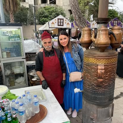 A woman and man standing together at an outdoor food stall with large traditional clay pots. The man wears a red apron and red hat, while the woman is dressed in a blue dress with a light-colored purse.