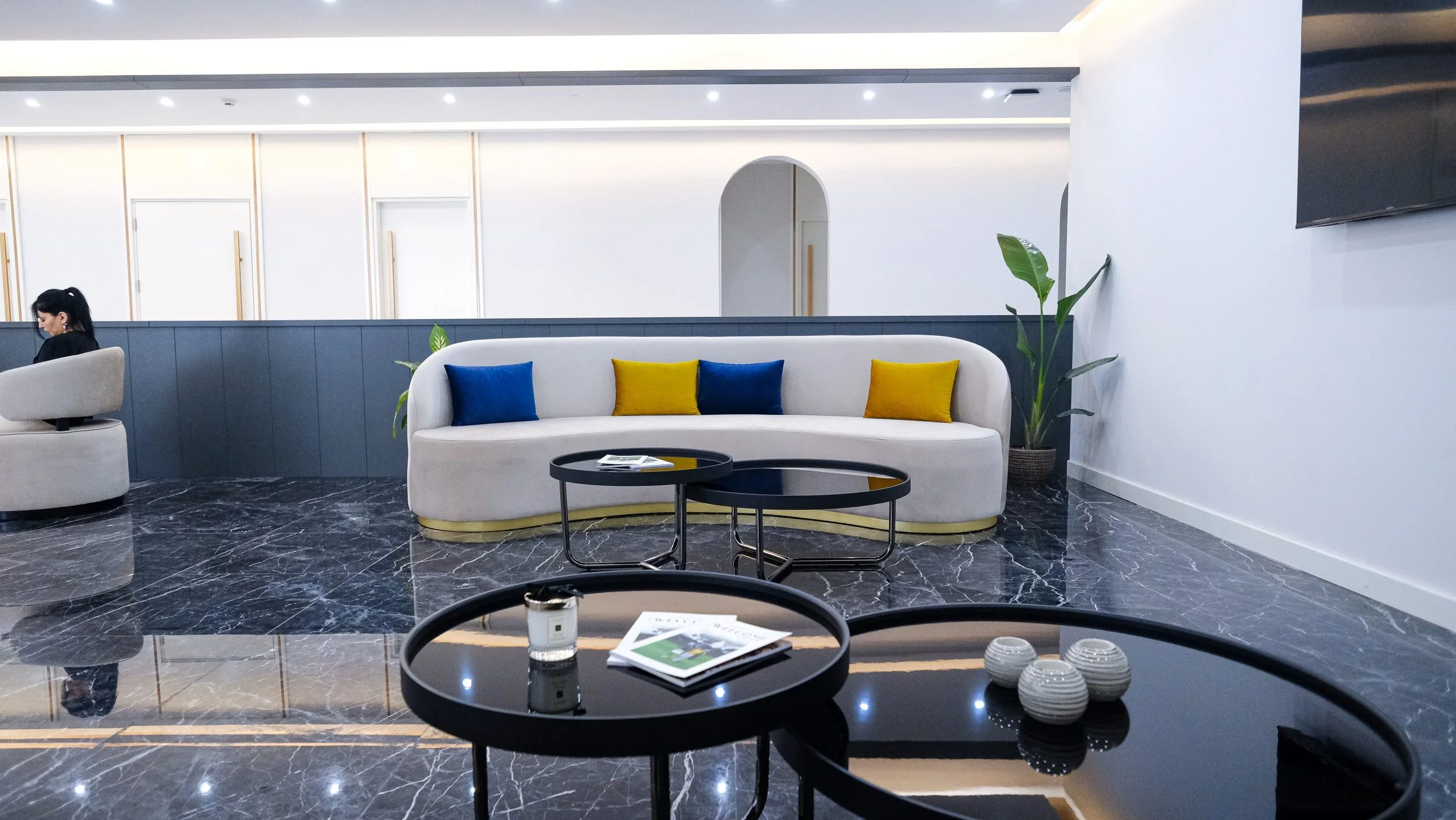 Modern hotel lobby with white curved sofa decorated with blue and yellow pillows, black marble coffee tables, a potted plant, and a large TV on the wall.