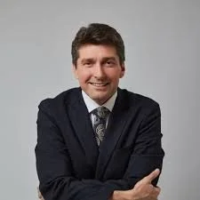A man in a dark suit with a tie, smiling and posing with crossed arms against a plain gray background.