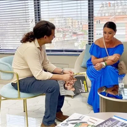 A man and a woman are sitting in a waiting area near large windows. The man is holding a magazine, and they are engaged in conversation. The woman is wearing a bright blue dress and appears to be listening.