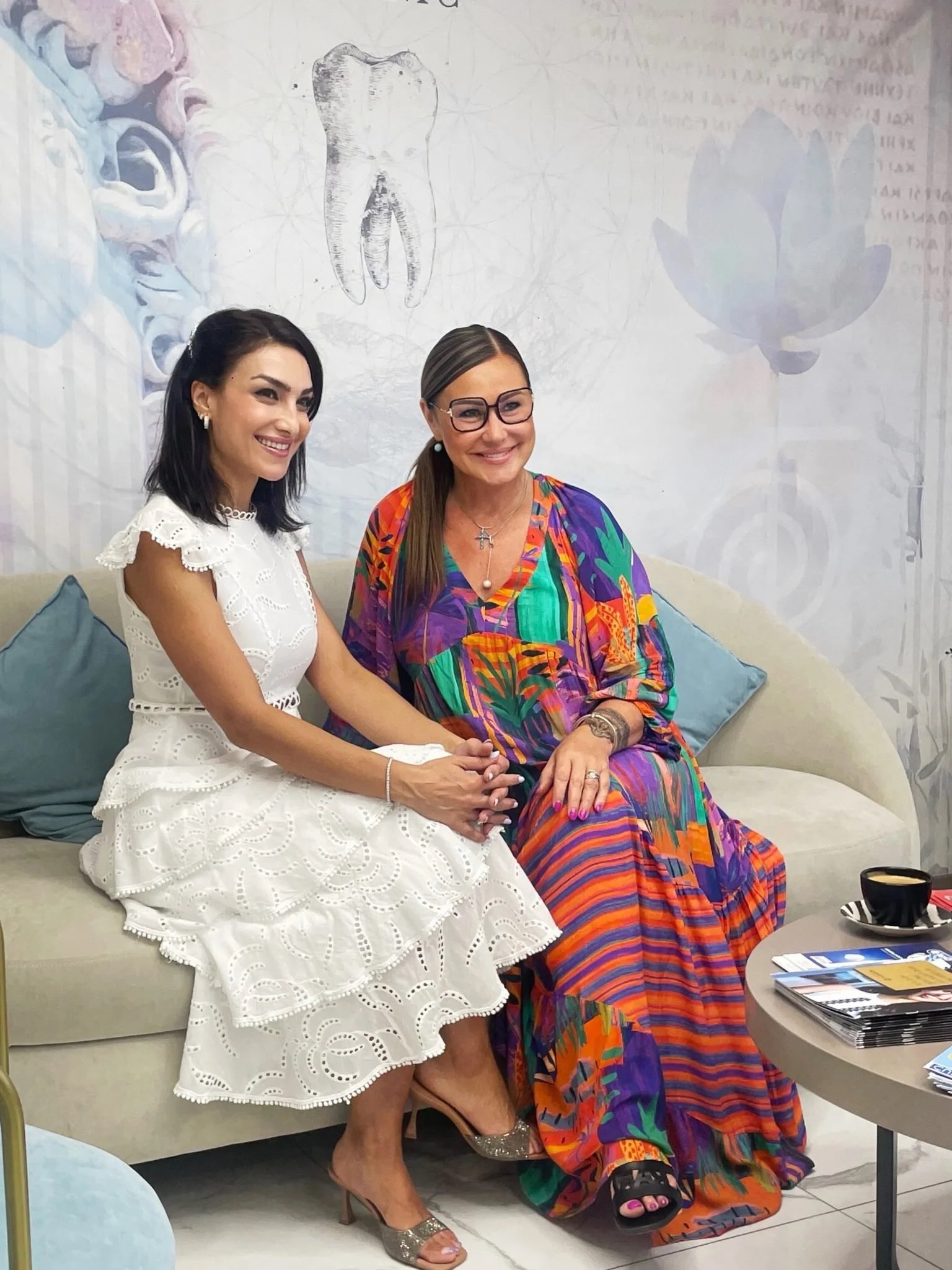 Two women sitting on a beige sofa, holding hands and smiling. The woman on the left is wearing a white dress with eyelet details and gold high heels. The woman on the right is wearing a colorful, patterned dress and glasses, with black slides. There are blue pillows behind them, and a round table with magazines and a black cup is in front of them. The background has wall art, including a tooth, a lotus flower, and a skull.