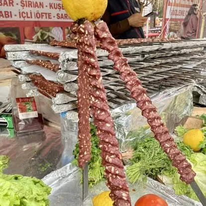 Three long, skewered pieces of raw meat with sesame seeds, topped with a lemon, displayed on a metal stand at a street food stall surrounded by lettuce, tomatoes, and other vegetables.