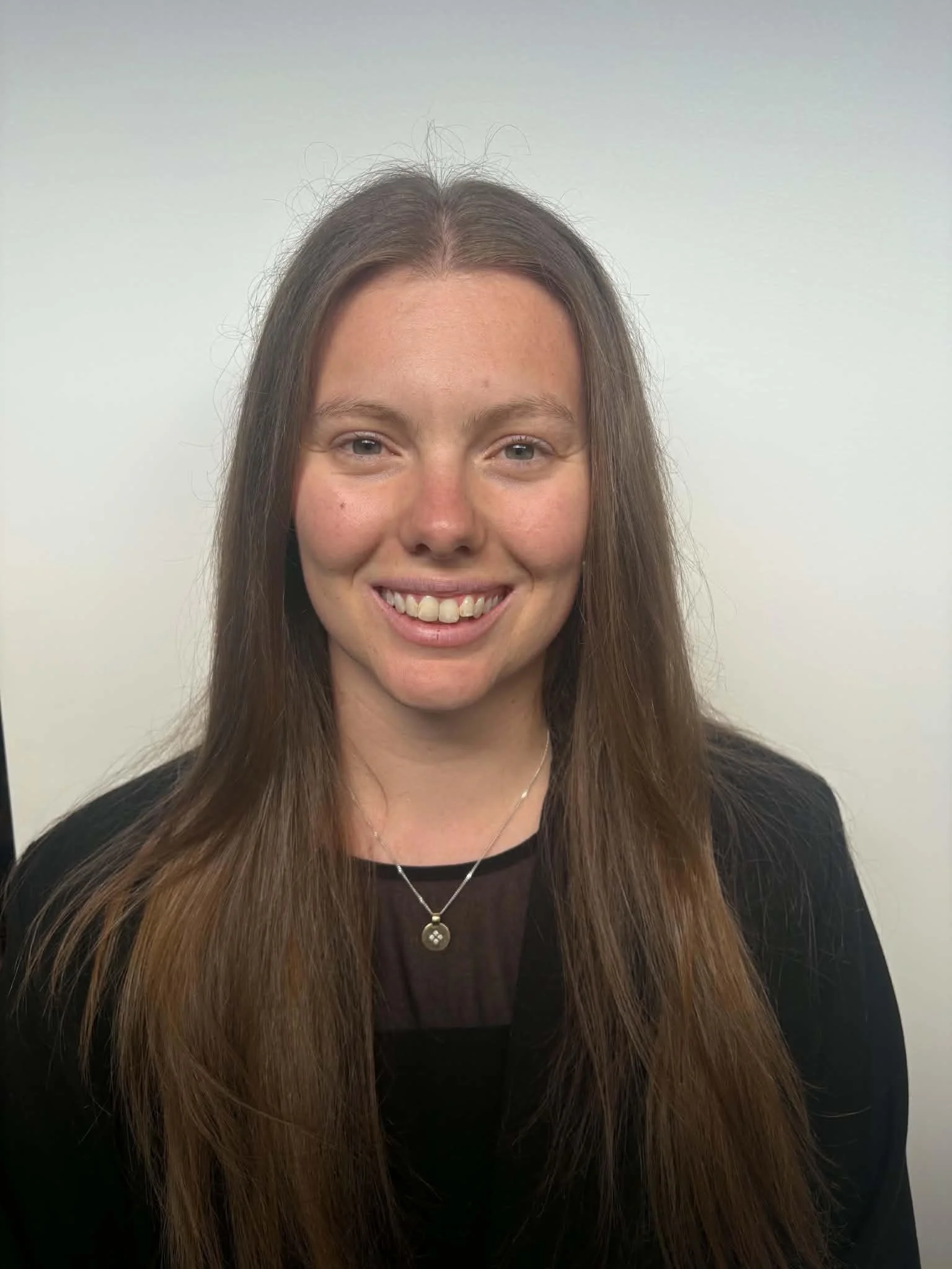 Close-up photo of a smiling speech pathologist with long brown hair wearing a black top, a necklace with a round pendant, and a subtle background.