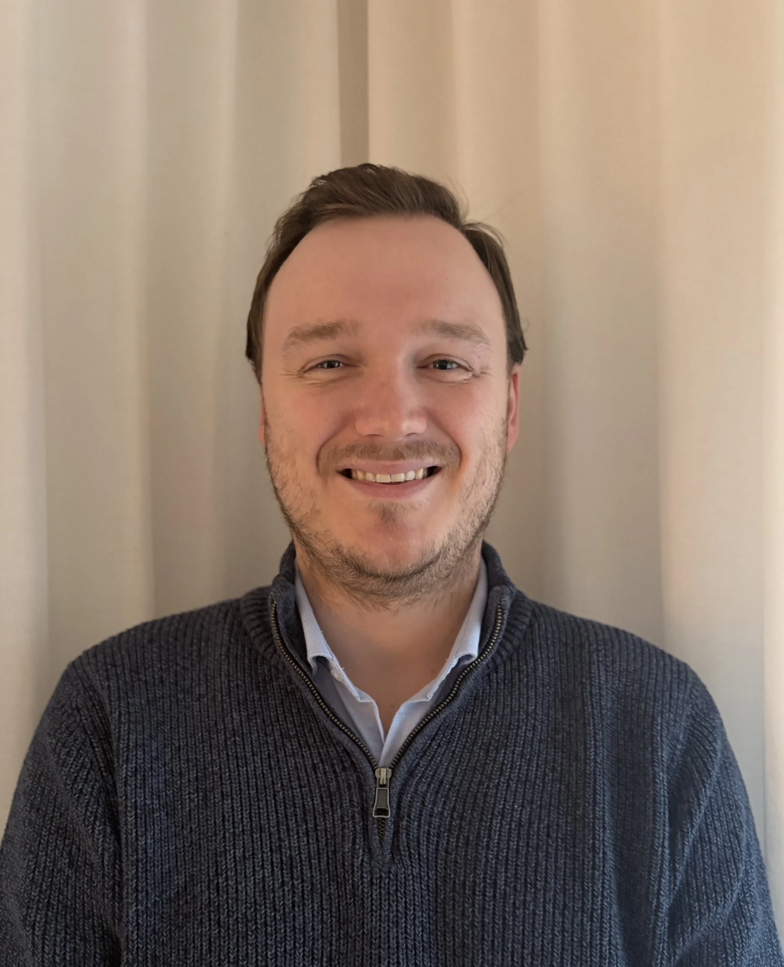 Close-up of a smiling psychologist who specialises in autism, ADHD and cognitive assessments in Bendigo, with short brown hair, light skin, wearing a dark zip-up sweater over a collared shirt, standing in front of beige curtains.